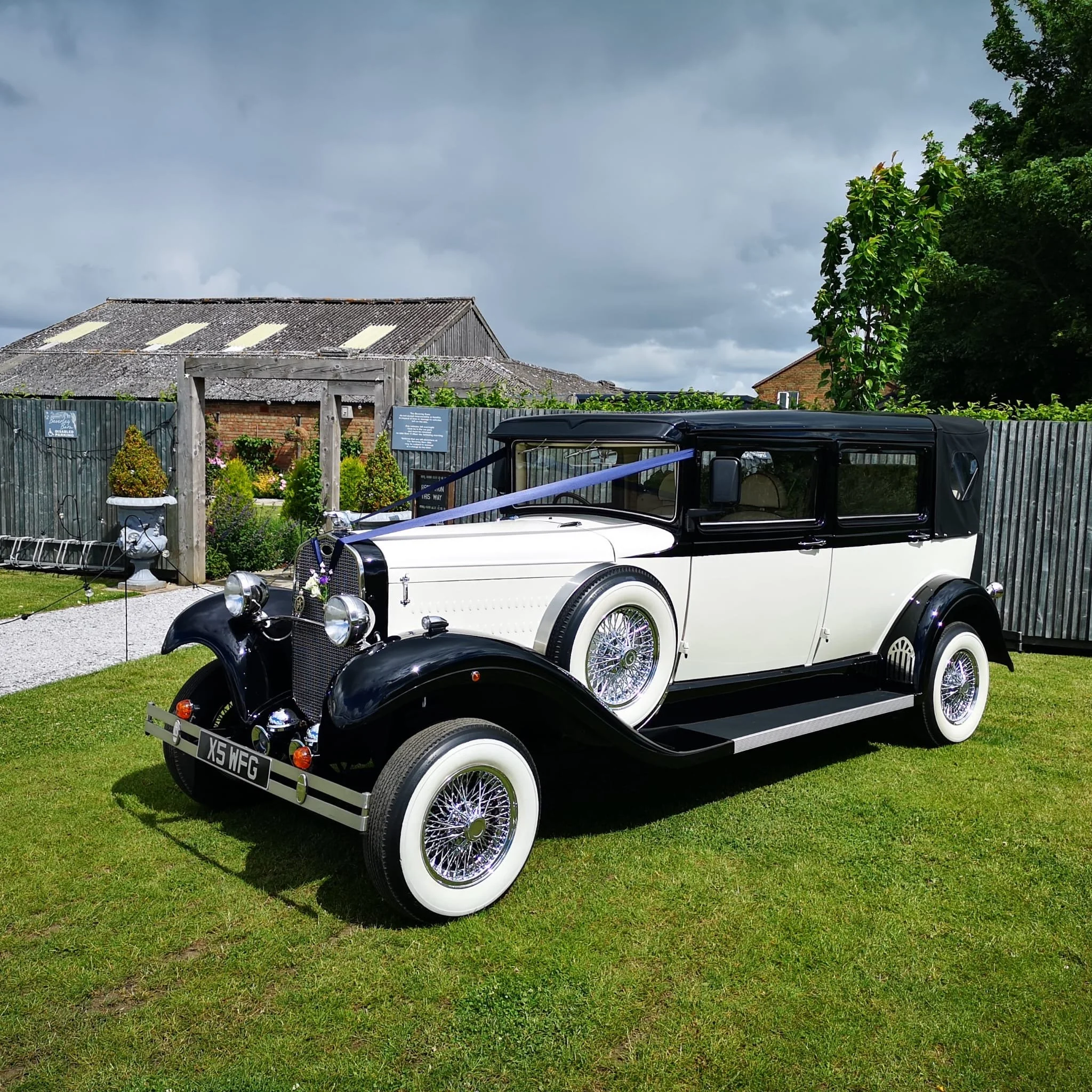 A vintage white and black automobile parked on a grassy area in front of a wooden fence, with a garden and a building with a weathered roofing under a cloudy sky.