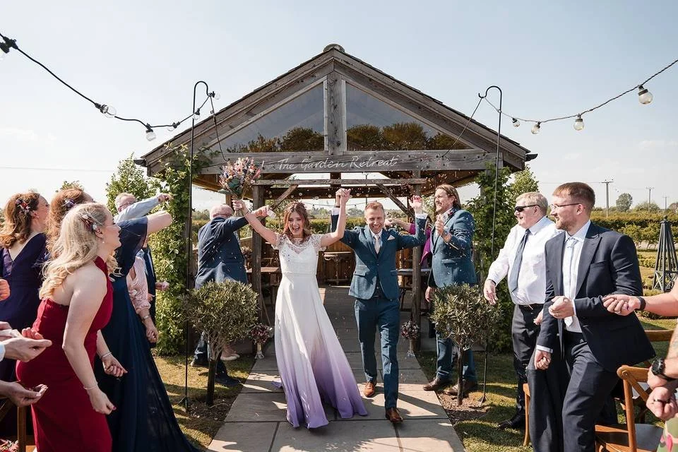Bride and groom celebrating with guests at an outdoor wedding ceremony, standing under an archway, with string lights overhead.