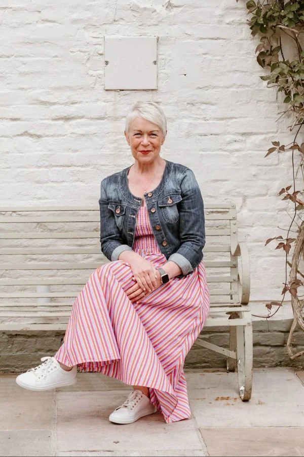 Older woman with short white hair sitting on a white wooden bench against a white brick wall, wearing a denim jacket, striped maxi dress, and white sneakers, with a smartwatch on her wrist.
