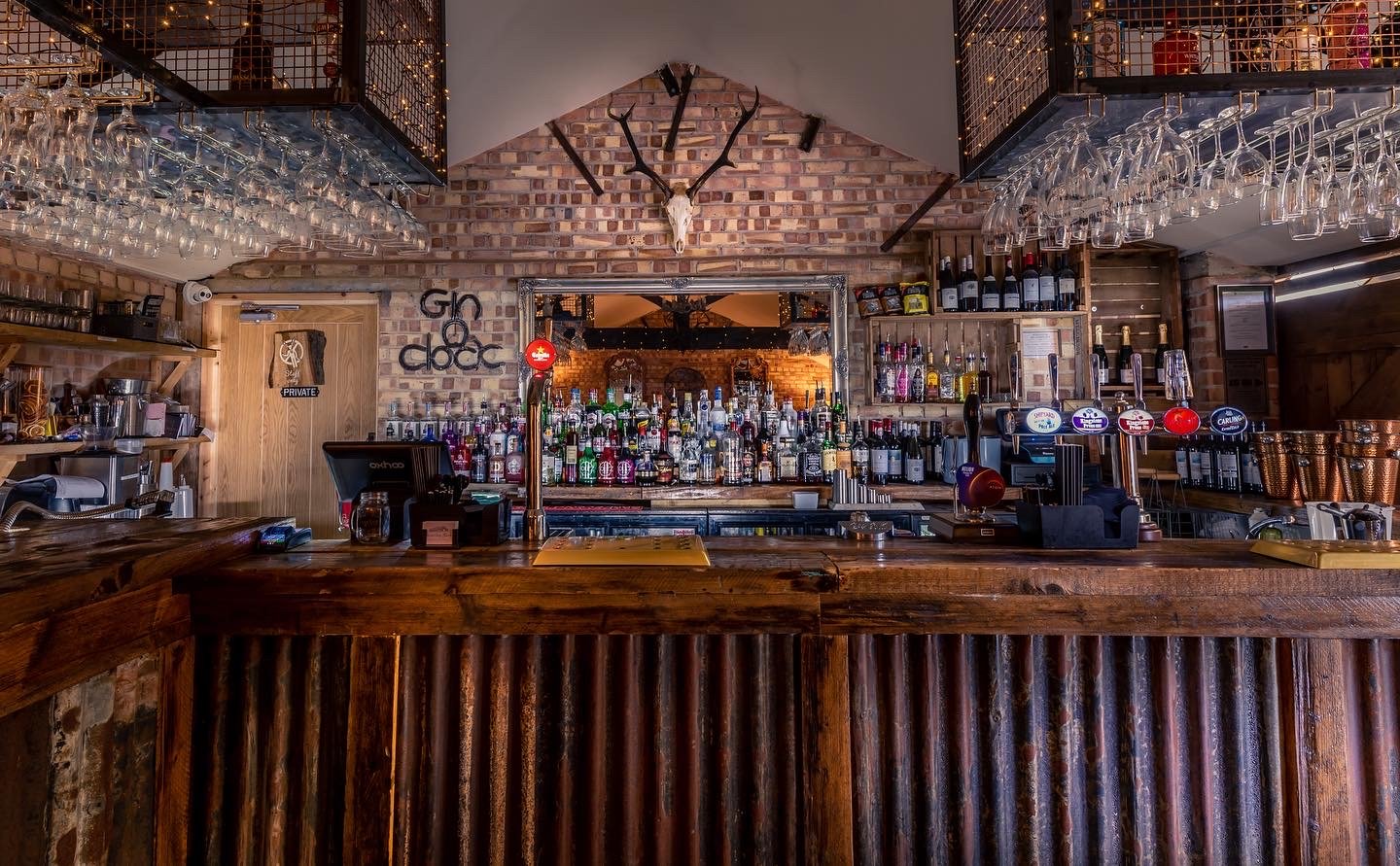 Interior of a bar with a rustic wooden counter, shelves filled with bottles of alcohol, hanging wine glasses, and brick walls. Decor includes a mounted animal skull with antlers and a modern clock. The setting appears warm and inviting.