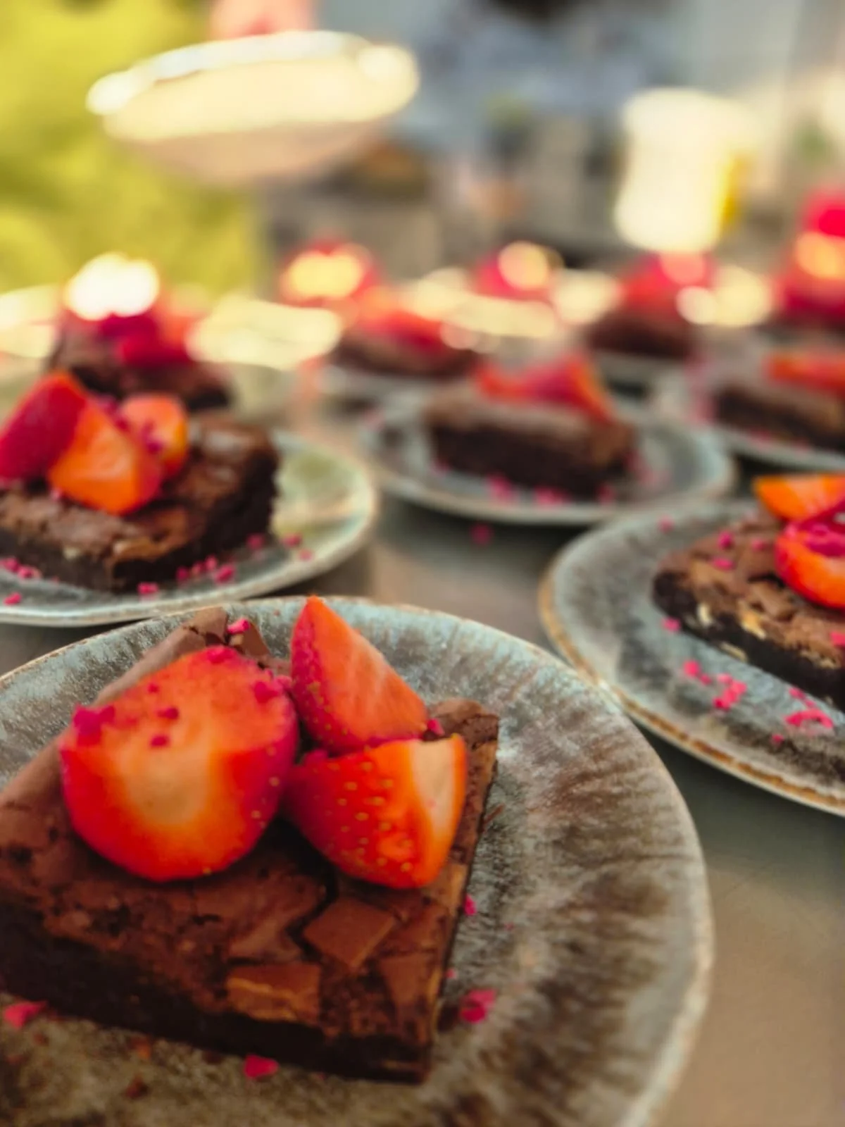 Close-up of chocolate brownies topped with sliced strawberries and pink sprinkles on a decorative plate.