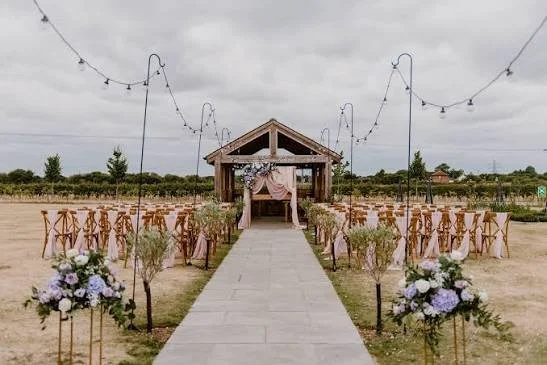Outdoor wedding ceremony setup with a wooden pavilion decorated with pink fabric, floral arrangements, and string lights on a cloudy day.