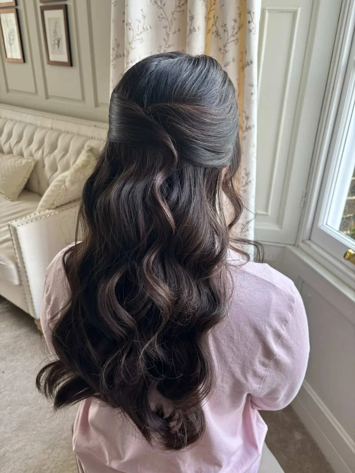 Back of a woman with long, dark wavy hair styled in loose curls, standing near a window with floral curtains in a room with beige decor.