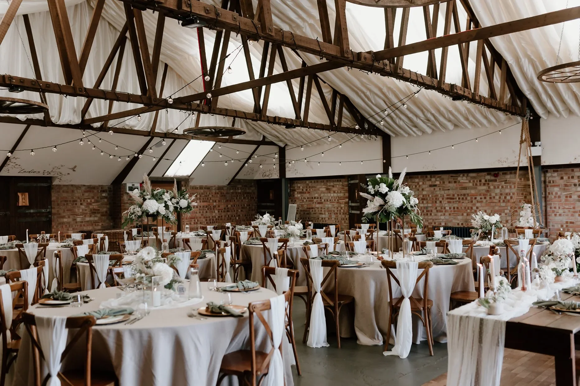Wedding reception setup in a rustic venue with round tables decorated with white tablecloths and floral centerpieces, surrounded by wooden chairs. The ceiling features wood beams and string lights, and the walls are brick with signs indicating 'Ladies' and 'Gents' restrooms.