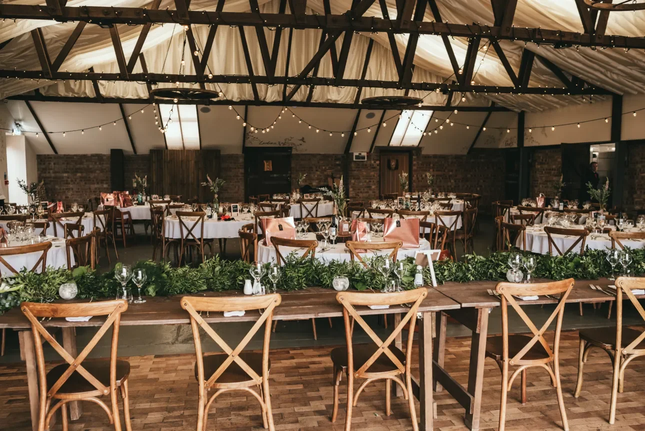 Elegant event space decorated with string lights, featuring round tables with white tablecloths, glassware, and floral centerpieces, along with a wooden table at the front adorned with greenery.