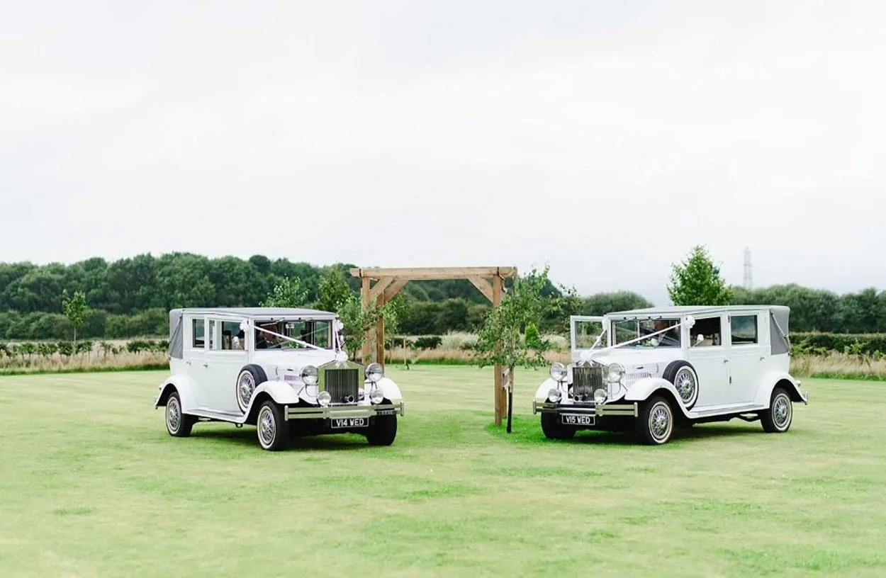 Two vintage white cars parked on a grassy field with a wooden wedding arch between them and small trees in the background.
