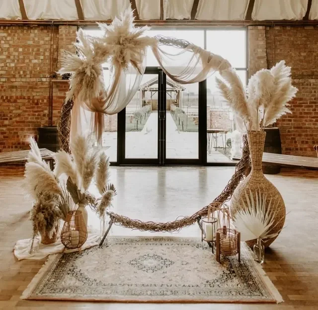 Bohemian-style indoor wedding or event backdrop with pampas grass arrangements in vases, a decorated arch with draped fabric, lanterns, and a rug on a wooden floor, with large glass doors and brick walls in the background.