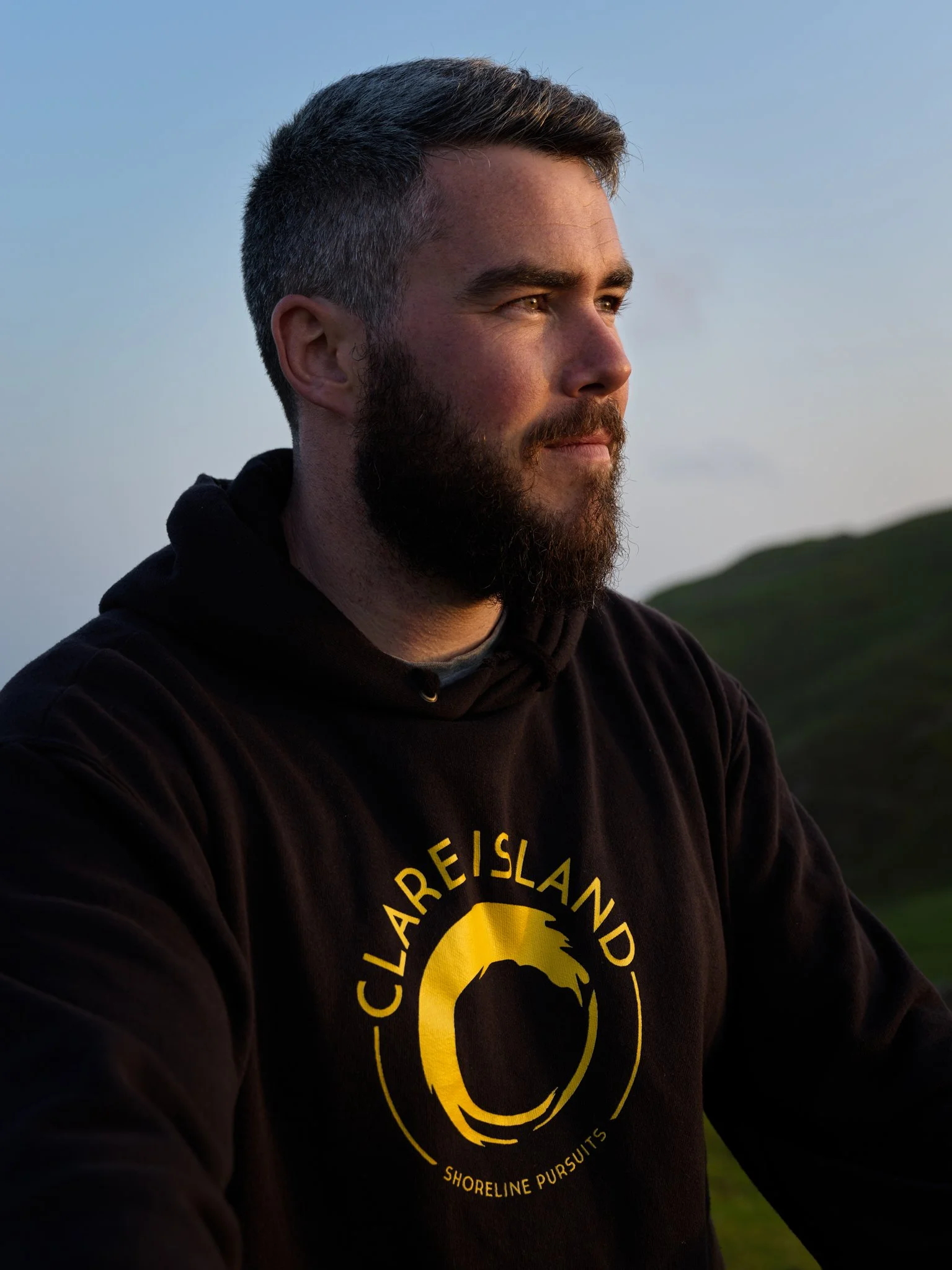 A man with short dark hair and a beard, wearing a black hoodie with a yellow circular logo reading "Clare Island" and "Shoreline Pursuits," standing outdoors during the evening with hills in the background.
