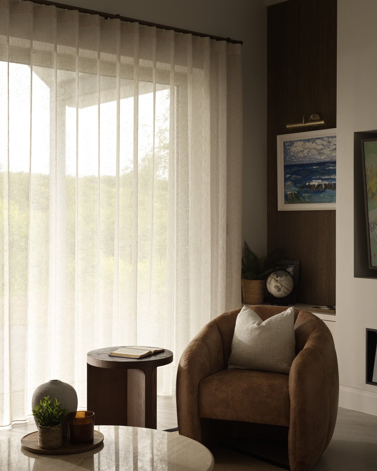 Cozy modern living room with a brown armchair, round side table, sheer curtains, indoor plants, artwork, and globe