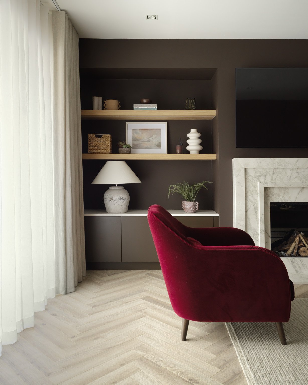 Living room with beige curtains, dark accent wall, built-in shelves with decor items, white fireplace with logs, red velvet armchair, off-white floor, and a narrow beige rug.