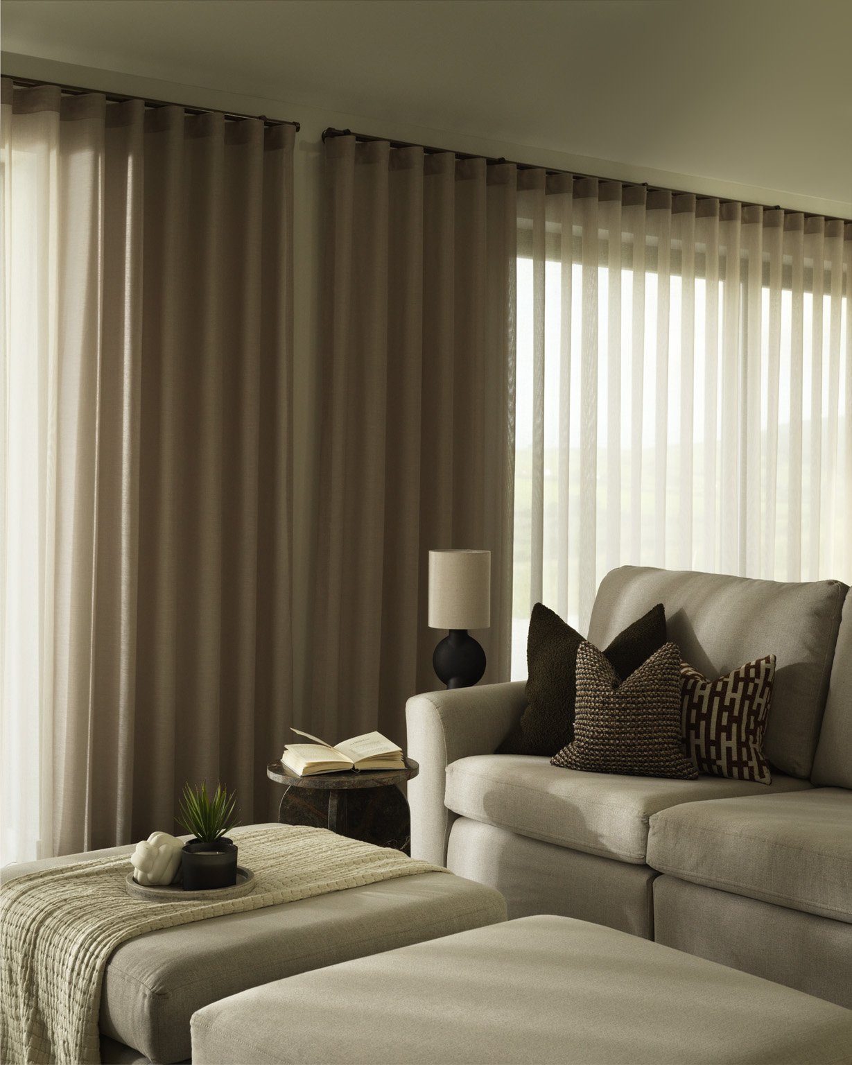 Living room with beige curtains, a beige sofa with black and patterned throw pillows, a side table with a lamp, a small table with an open book and decorative items, and large windows letting in natural light.