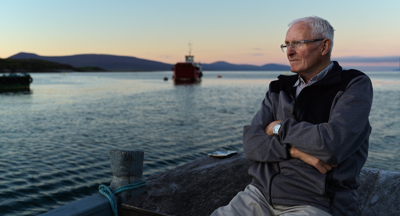 An older man with gray hair, glasses, and a watch, sitting by the water on a dock at sunset, with hills in the background and boats in the distance.