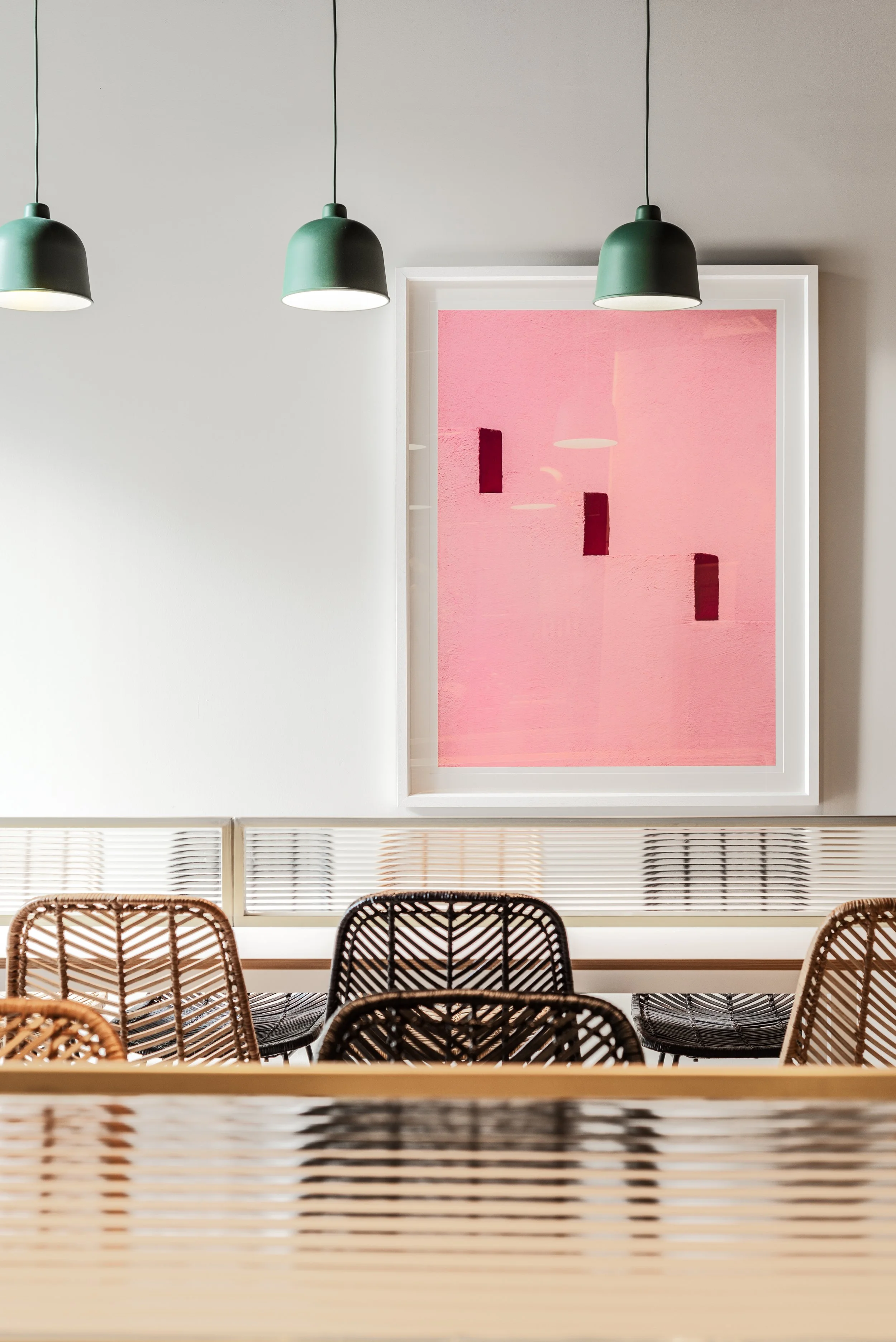 Interior of a modern space with white walls, three green pendant lights hanging from the ceiling, a pink abstract artwork on the wall, and wicker chairs arranged around a table.