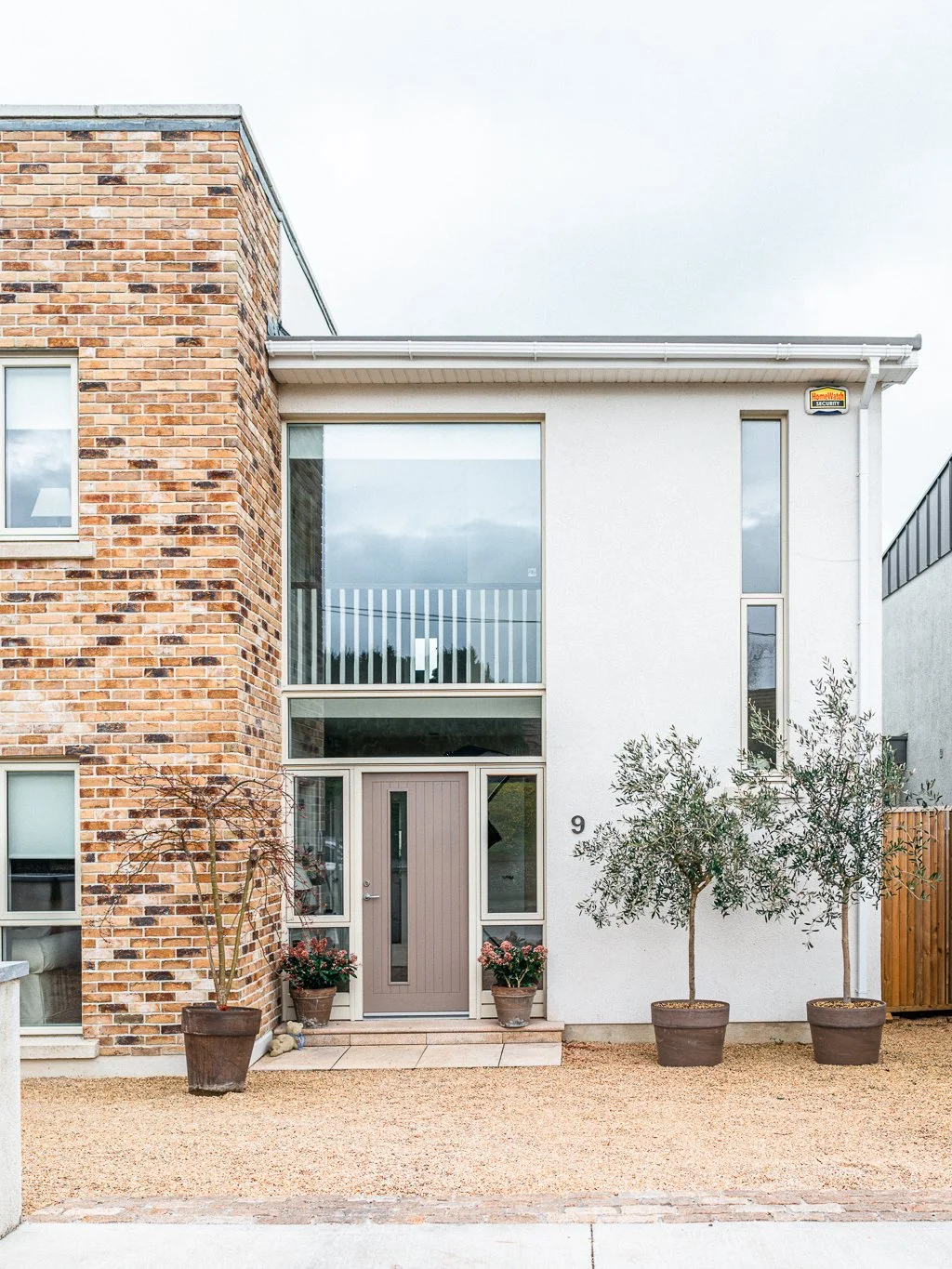 Modern house with a brick and white exterior, large windows, potted plants, and small trees in the front yard.