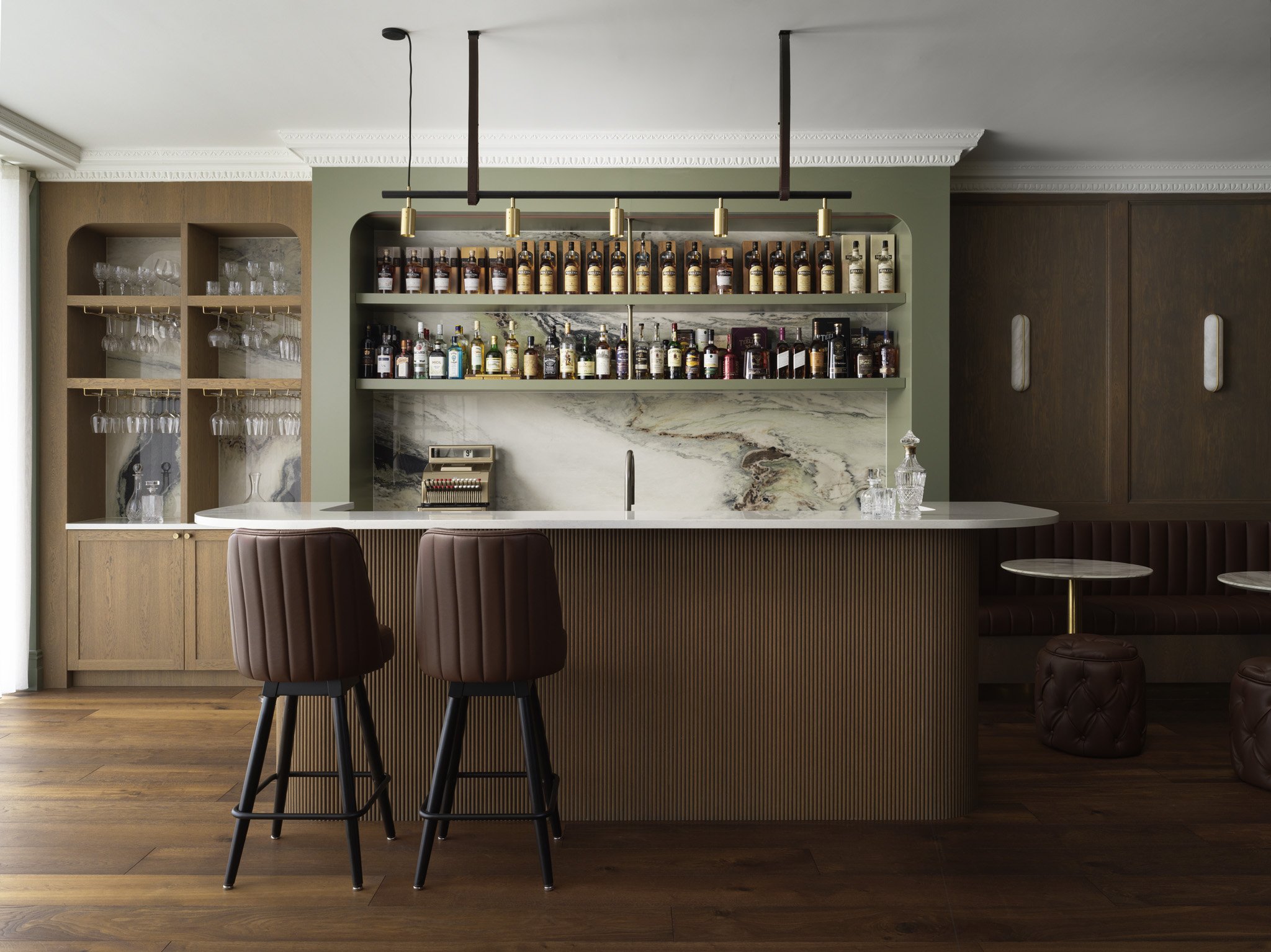 Bar area with wooden and marble finishes, two brown upholstered bar stools, shelves with bottles and glassware, and a marble countertop.