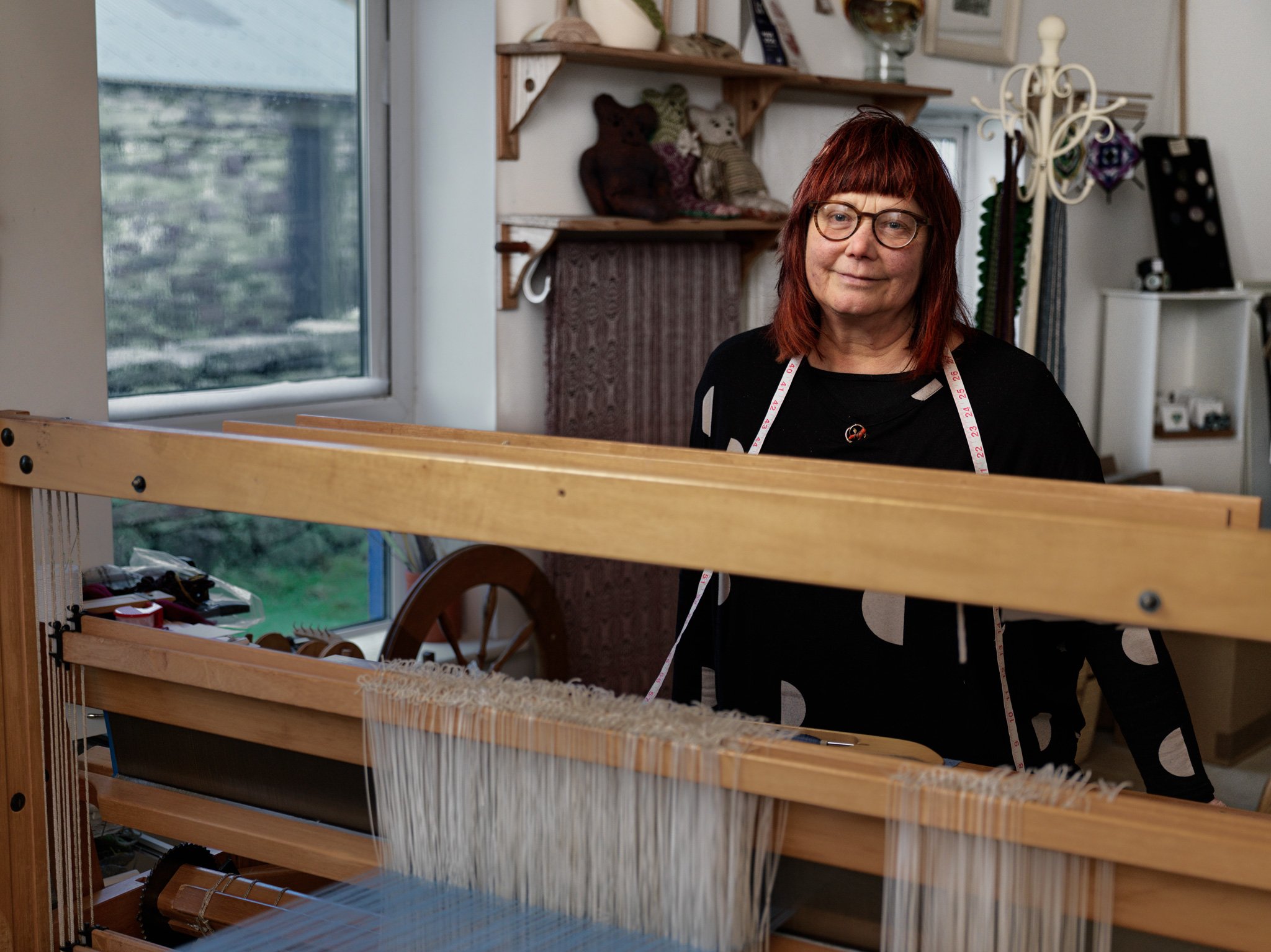 A woman with glasses and red hair standing behind a weaving loom in a cozy room with shelves of yarn and stuffed bears, near a window with a view of greenery outside.