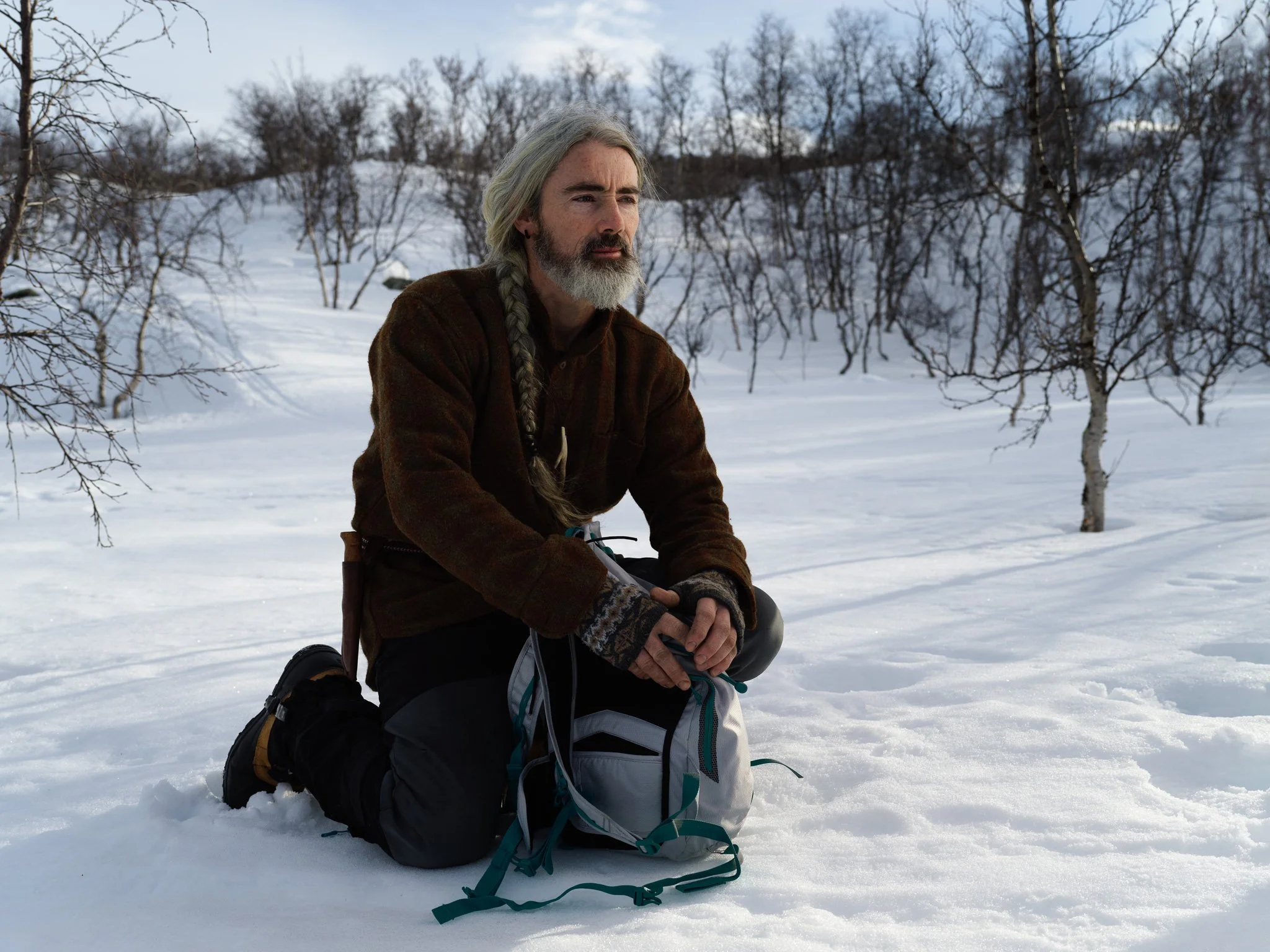 A man with gray hair and beard kneels in the snow with a backpack, surrounded by leafless trees, during winter.