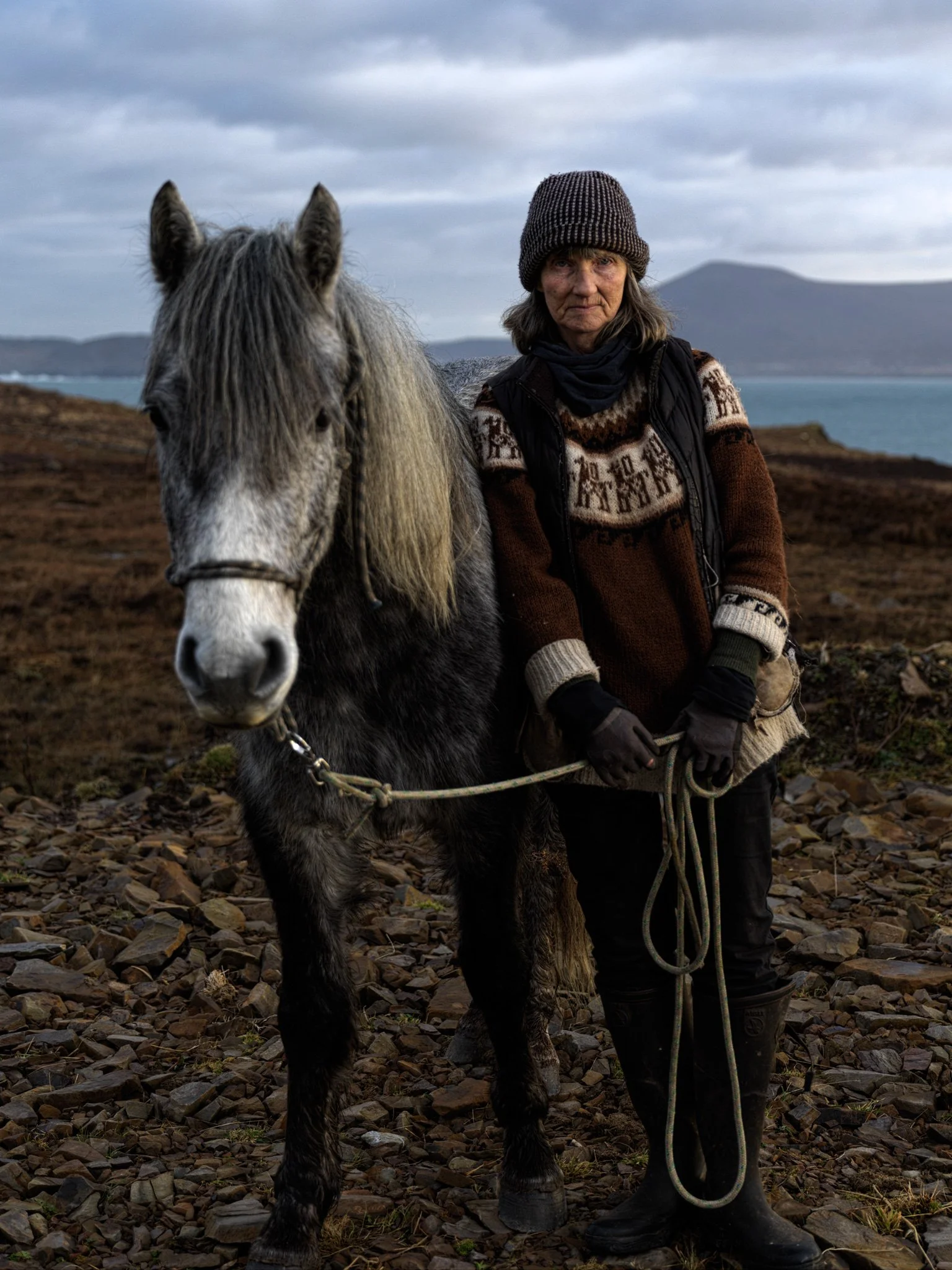 A woman standing next to a gray and black horse on rocky terrain, with water and hills in the background, under cloudy skies.
