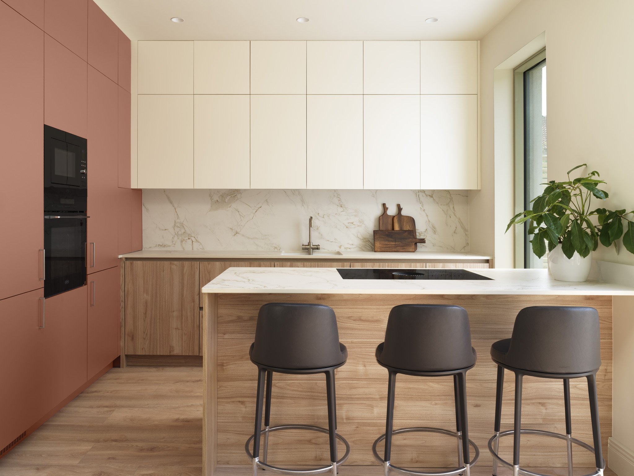 Modern kitchen with pink and beige cabinetry, marble backsplash, wooden accents, a marble island with three black barstools, and a potted plant by the window.