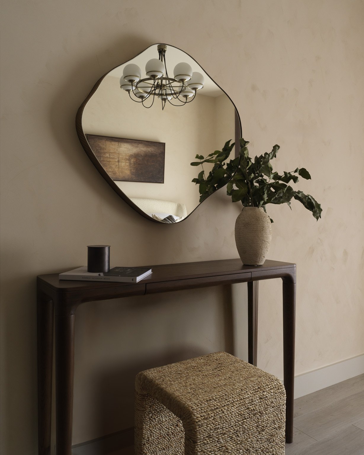 A decorative living space with a wall mirror, a vase with green plants on a wooden table, a candle, jewelry, a stool, and a chandelier reflected in the mirror.