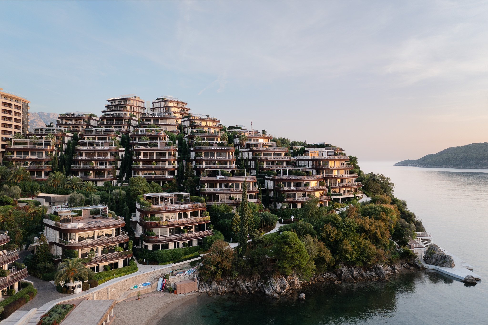 Luxury hillside Hotel Apartment complex Dukley Gardens  overlooking a body of water, with a small beach and greenery in the foreground, and hills in the distance at sunset.