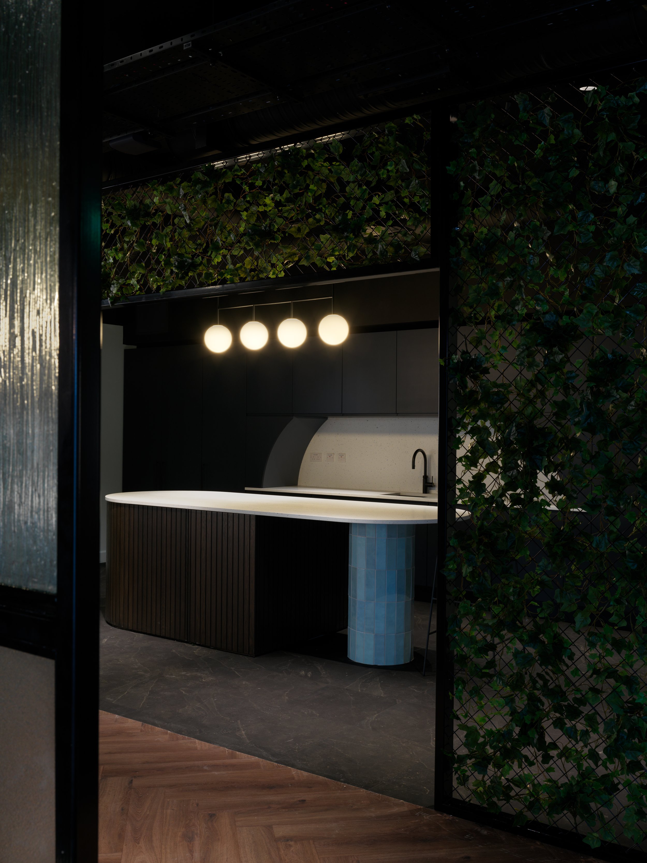 Modern kitchen with black cabinetry, a white countertop, black sink faucet, three round pendant lights, and a partial view of a wooden floor and green leafy wall.