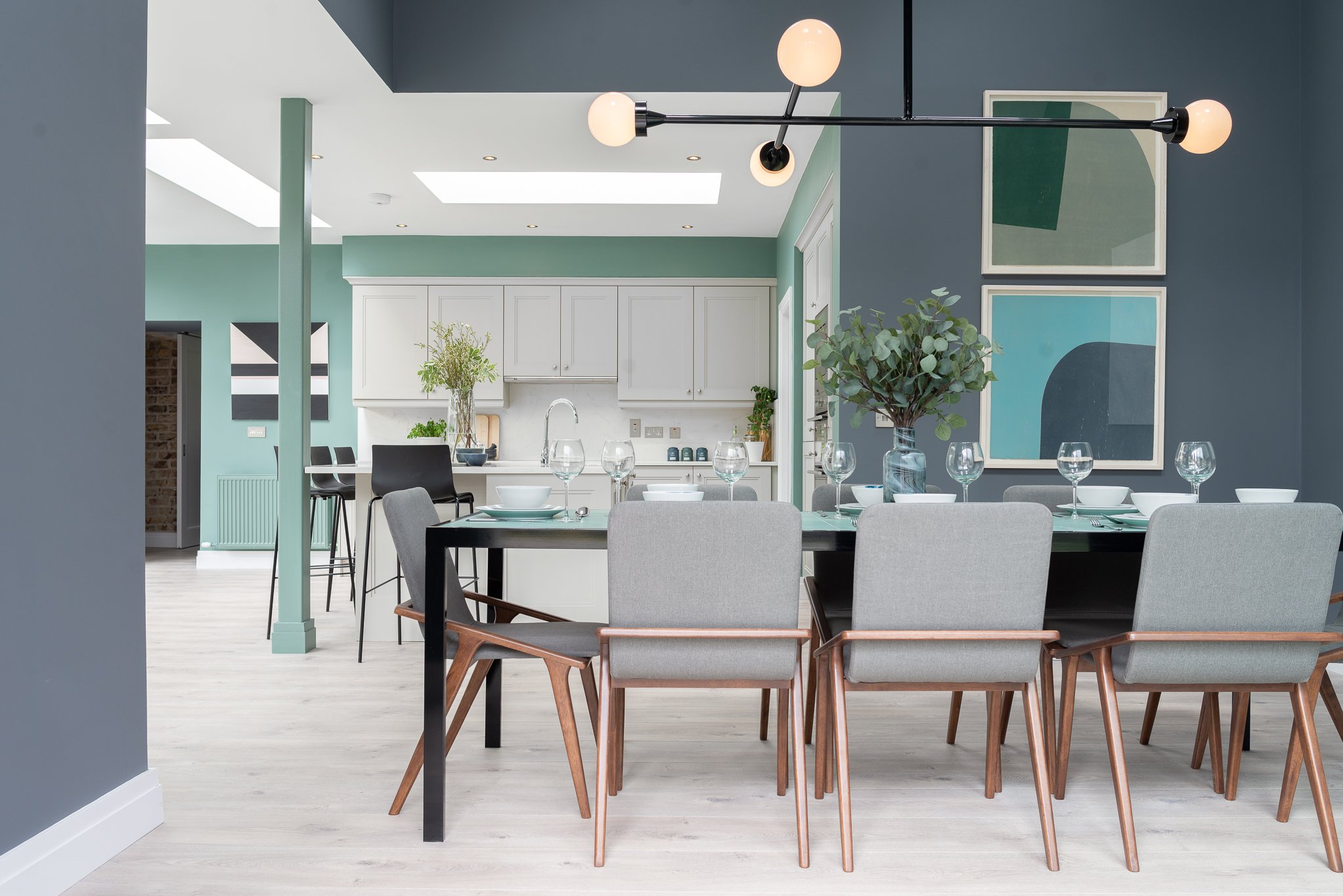 Modern dining room with a black table, gray chairs, and glassware, situated adjacent to a kitchen featuring white cabinets and minimalist decor. Artistic wall art and a large vase with greenery are also visible.