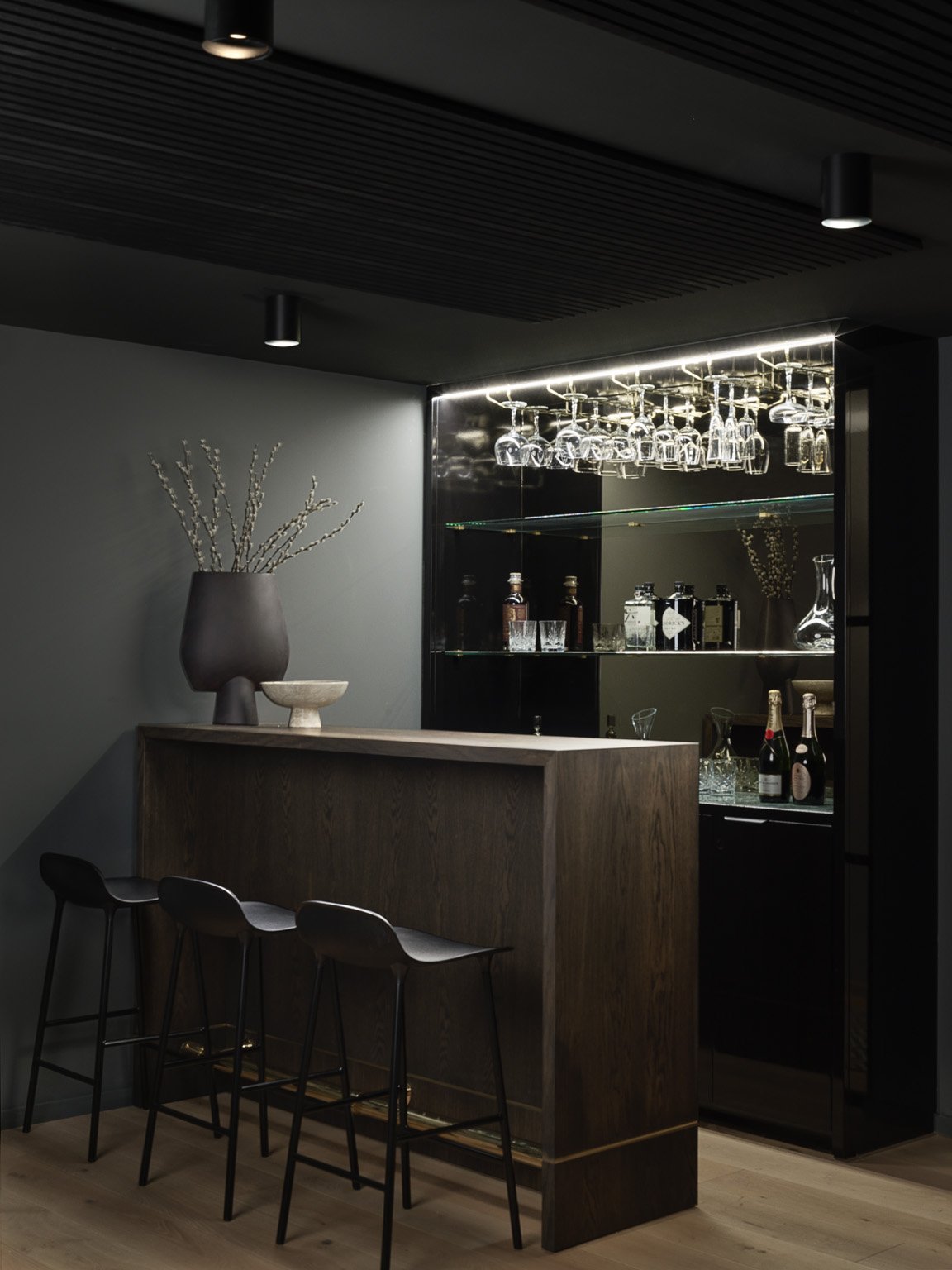 Modern bar area with a dark wooden counter, black chairs, and glassware hanging above. A large gray vase with branches and a small stone bowl are on the counter, and a mirrored cabinet with bottles and glasses is in the background.