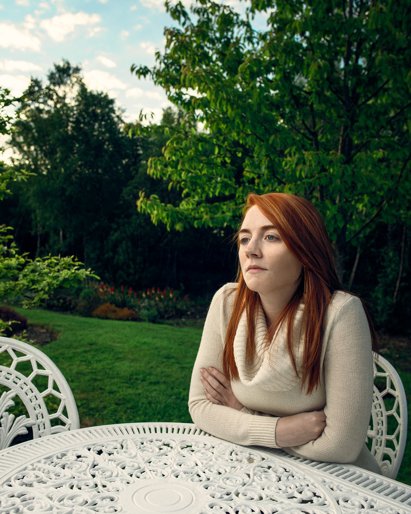 A woman with red hair and a beige sweater sitting at a white ornate outdoor table in a garden with green trees and plants, under a partly cloudy sky.