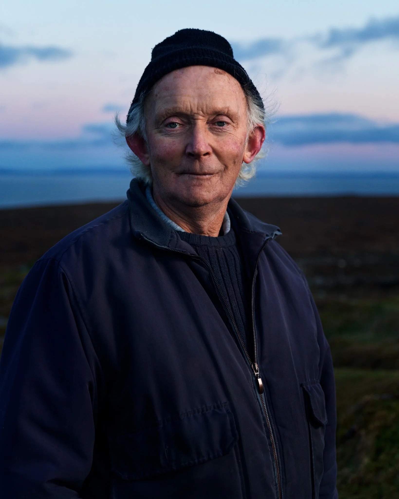 A portrait of an elderly man with gray hair and a beard, wearing a black beanie, navy jacket, and a dark sweater, standing outdoors during the early evening with a cloudy sky in the background.