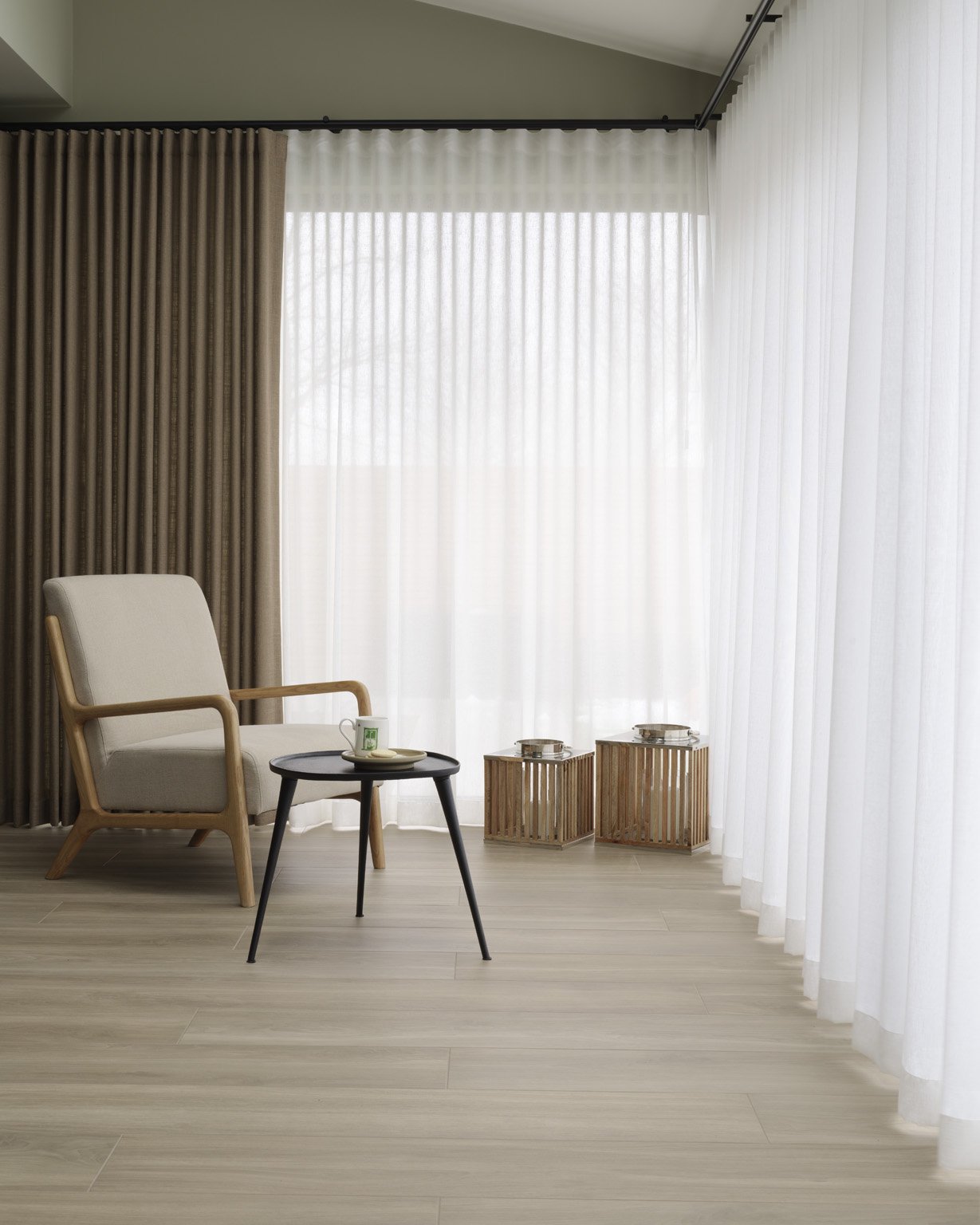 A minimalist living room corner with a white armchair, a small black side table with a white cup and saucer, and two wooden latticed side tables with covered trays on top, next to large sheer white curtains covering a window.