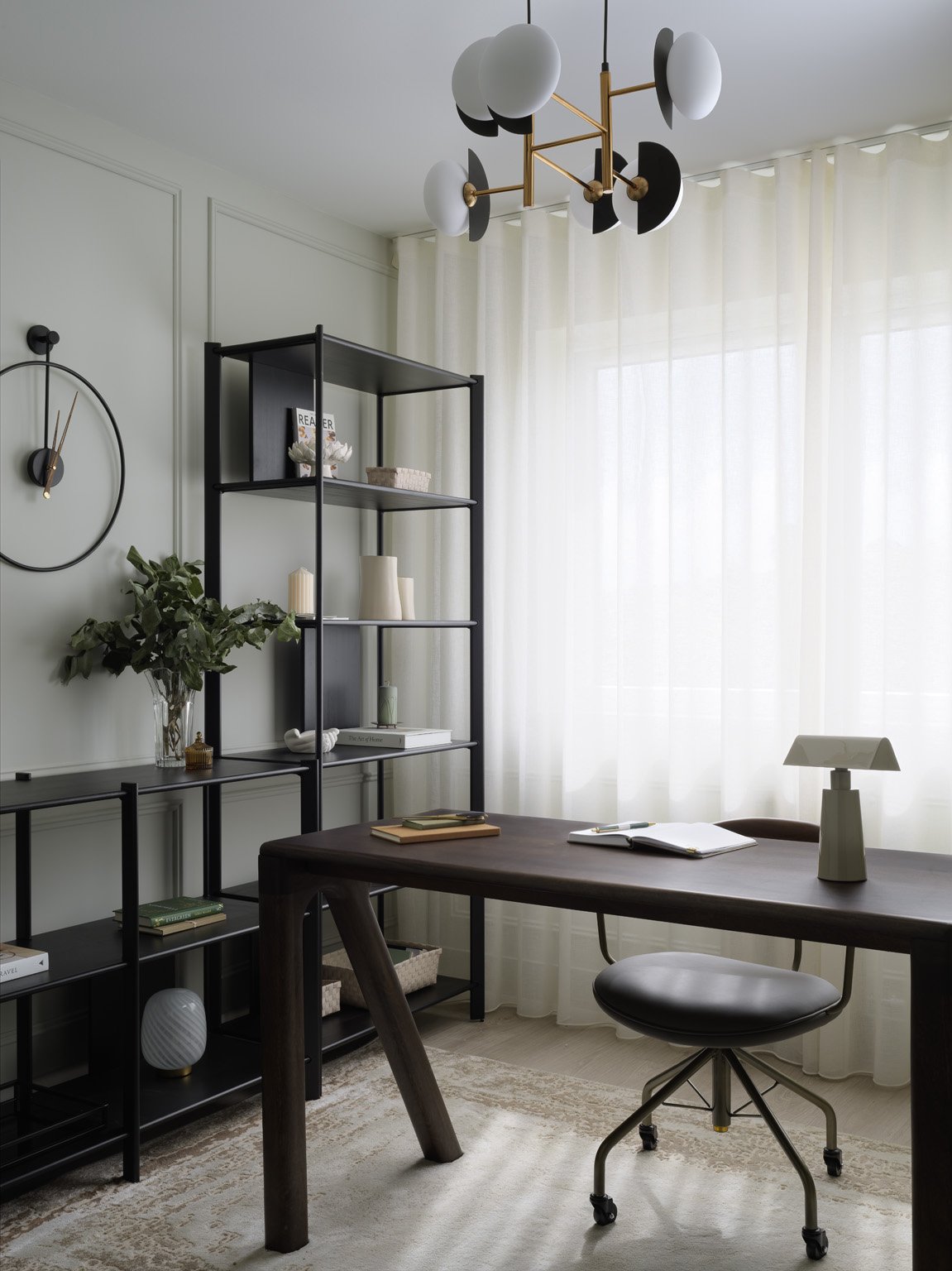 A modern home office with a dark wooden desk, black rolling chair, and a black shelving unit with books and decorative items. Light-colored curtains cover a large window, and a contemporary chandelier hangs from the ceiling.