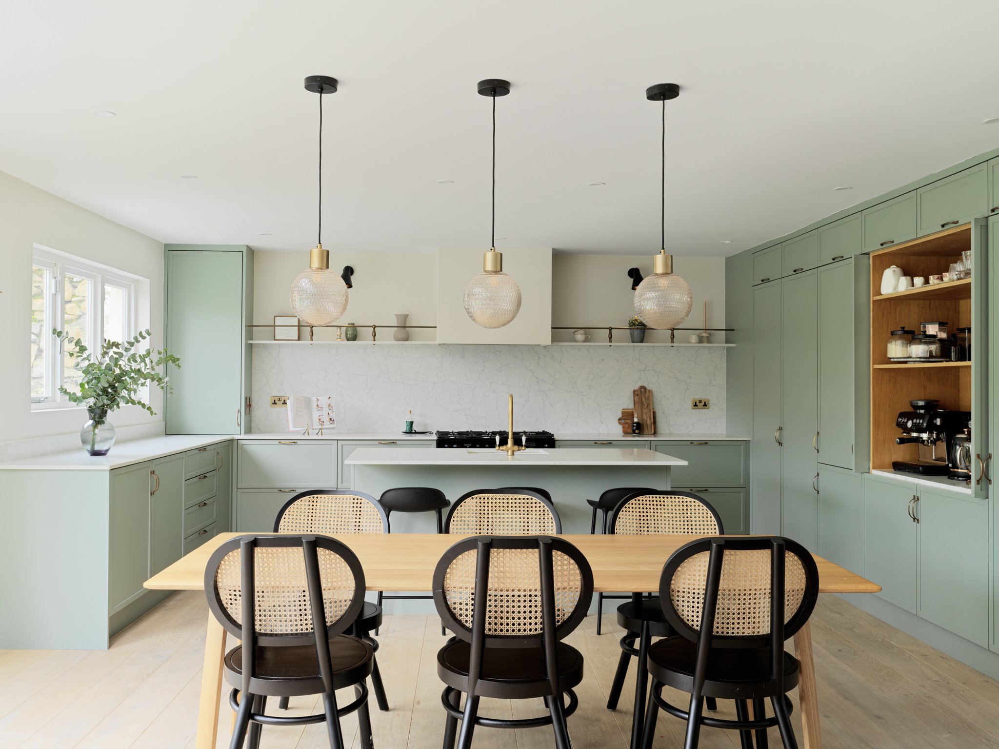 Modern kitchen with pastel green cabinets, a wooden dining table with six black chairs, pendant lights, a window with a plant, open shelving, and a white marble backsplash.