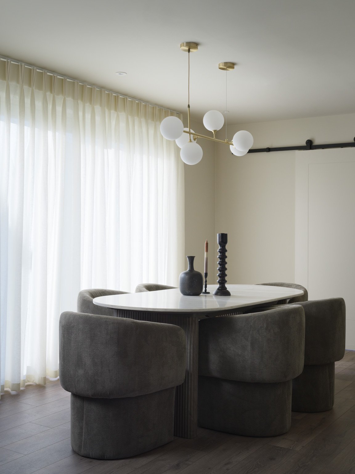 Modern dining room with a white oval table and six gray upholstered chairs. Light sheer curtains cover large windows, and a unique ceiling light fixture with six spherical bulbs hangs above the table. The table is decorated with three vases and a can