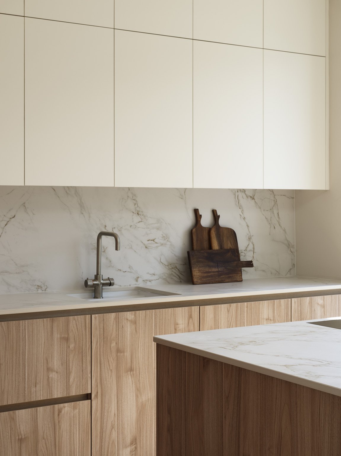 Modern kitchen with wooden cabinets, white marble countertop, a sink with a silver faucet, and two wooden cutting boards on the backsplash.