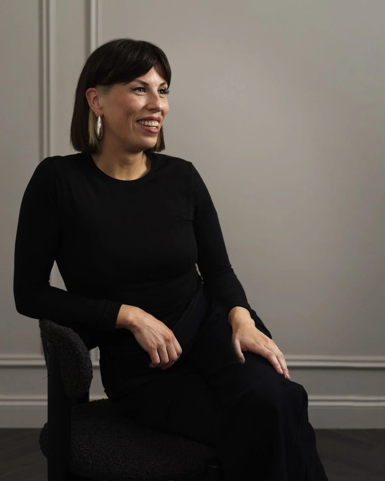 A woman with short dark hair, wearing a black top and earrings, sitting on a black chair against a plain light-colored wall, smiling and looking to her left.