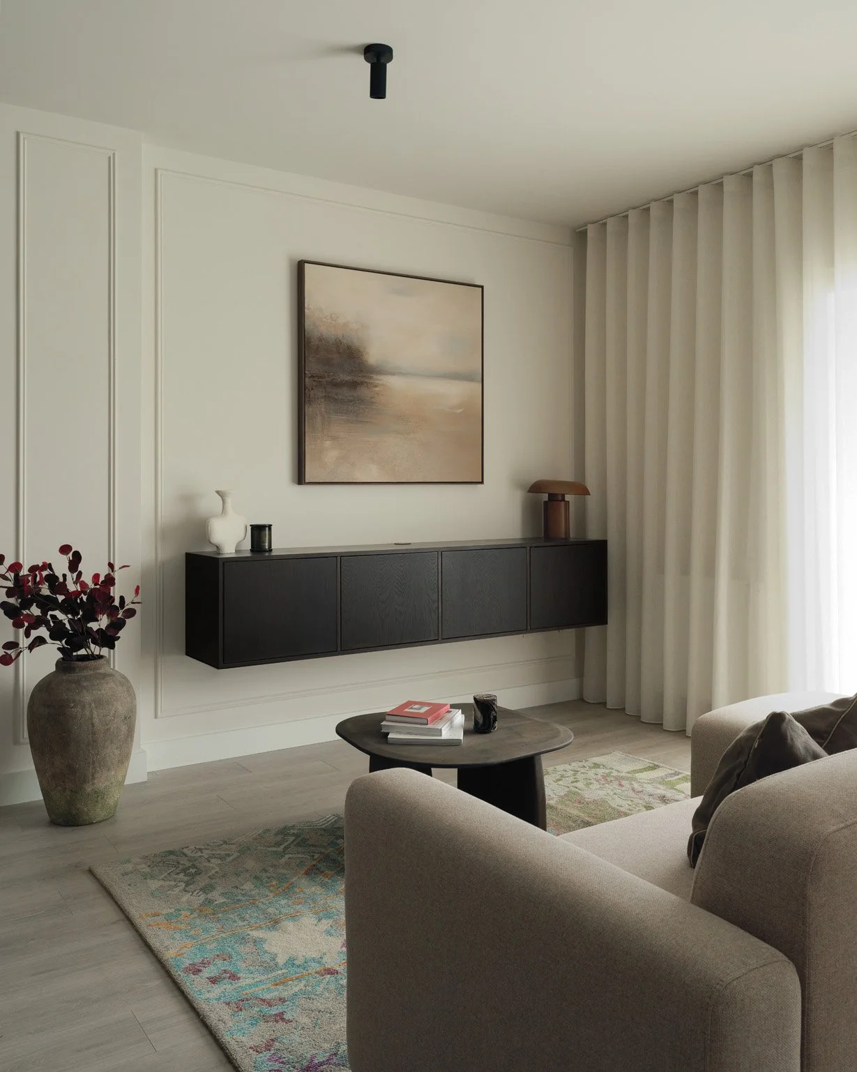 Living room with beige sofa, dark wood sideboard, beige curtains, abstract landscape painting, round coffee table with books and a mug, potted plant, and a ceiling light fixture.
