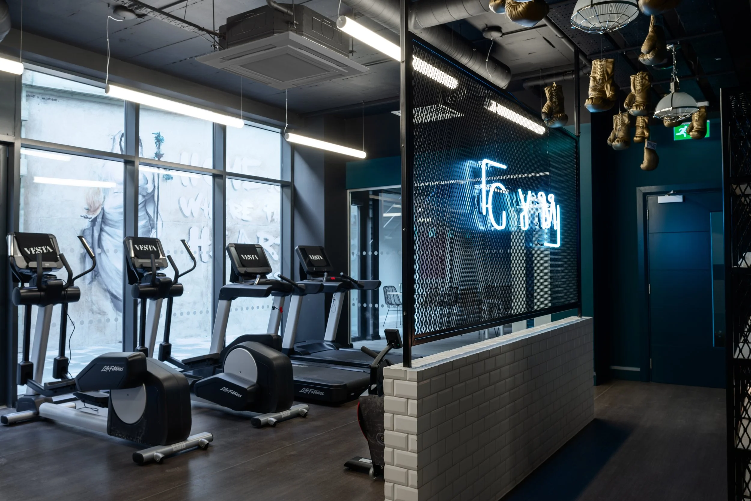 Modern gym interior with stationary bikes and a treadmill, large windows, and a neon sign with Asian characters, separated by a mesh divider with boxing gloves hanging above.