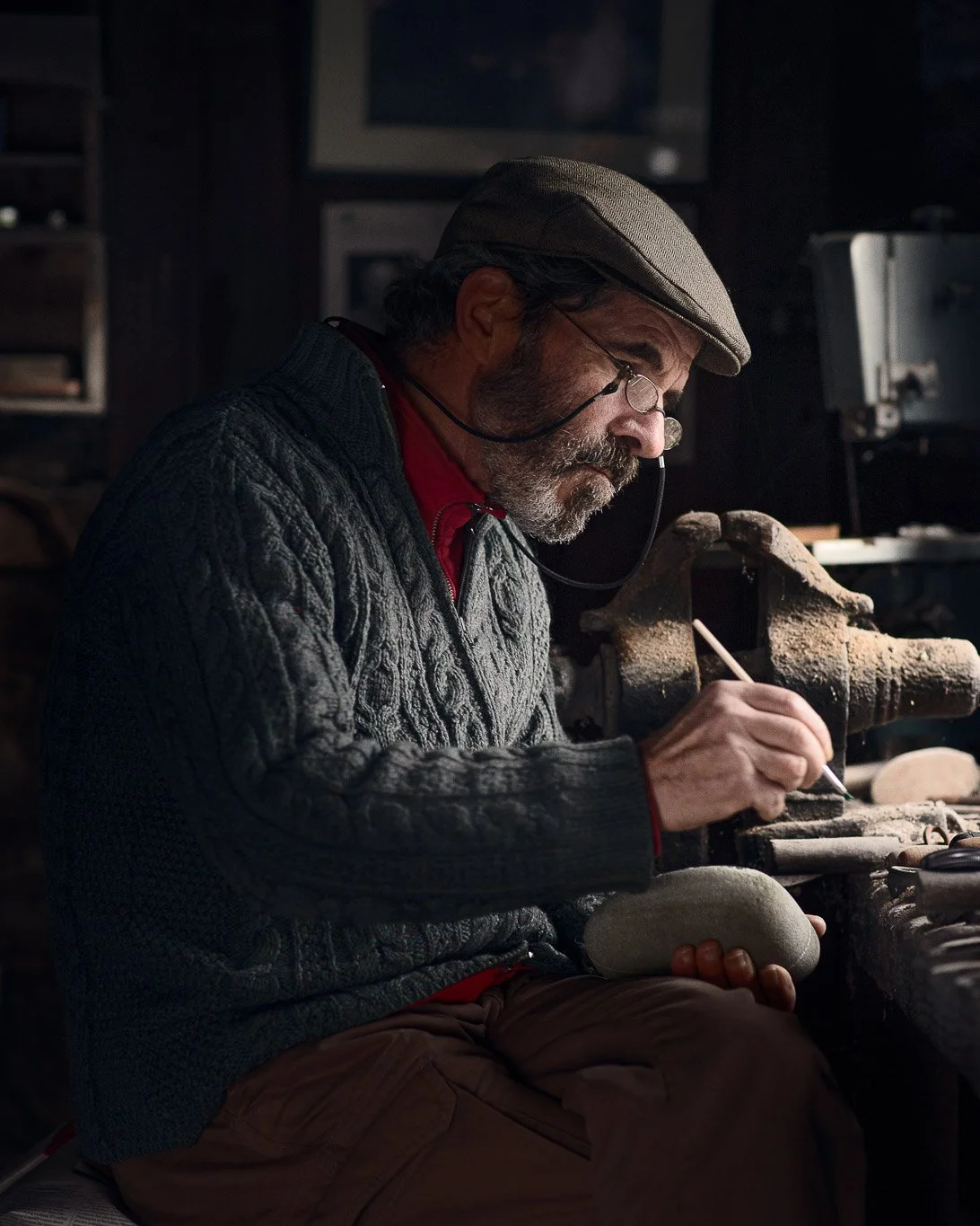 An elderly man with glasses, a cap, and a gray cable-knit sweater working on a stone carving at a workbench in a workshop.