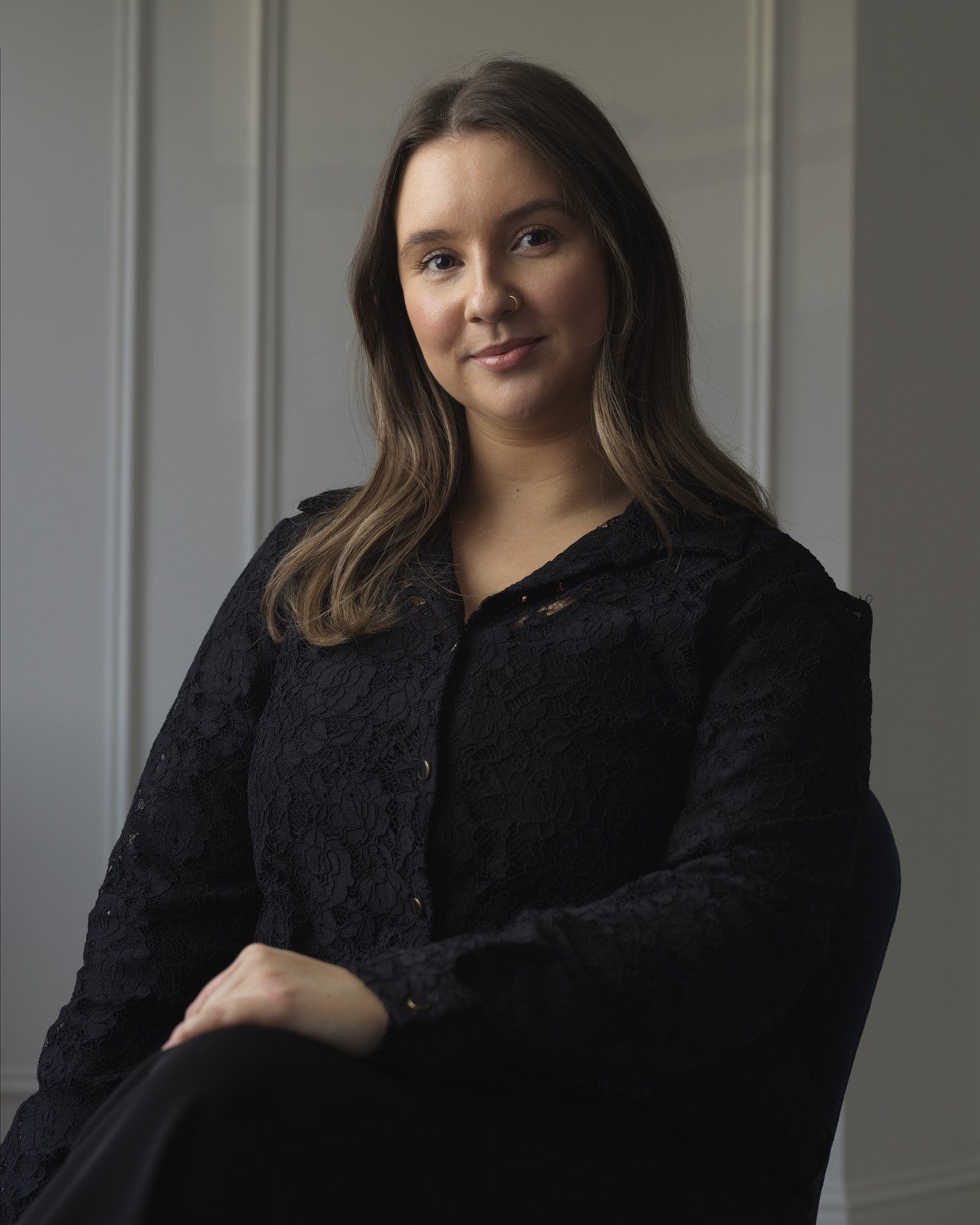 A young woman with brown hair and a nose piercing sitting in front of a plain background.