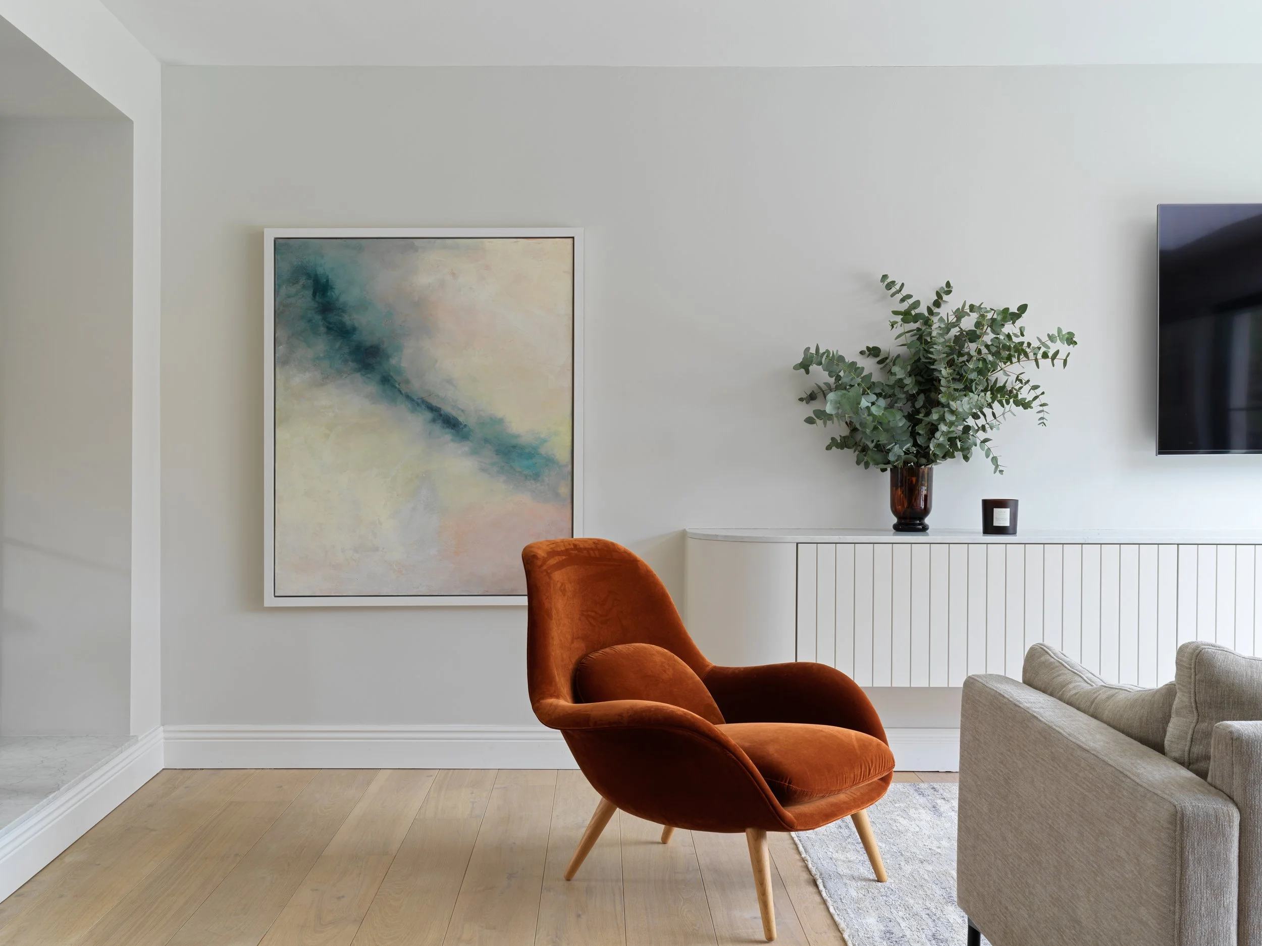 Living room with white walls, modern art painting, a rust-colored velvet armchair, a beige sofa, a white cabinet, a large plant in a dark vase, and a flat-screen TV mounted on the wall.