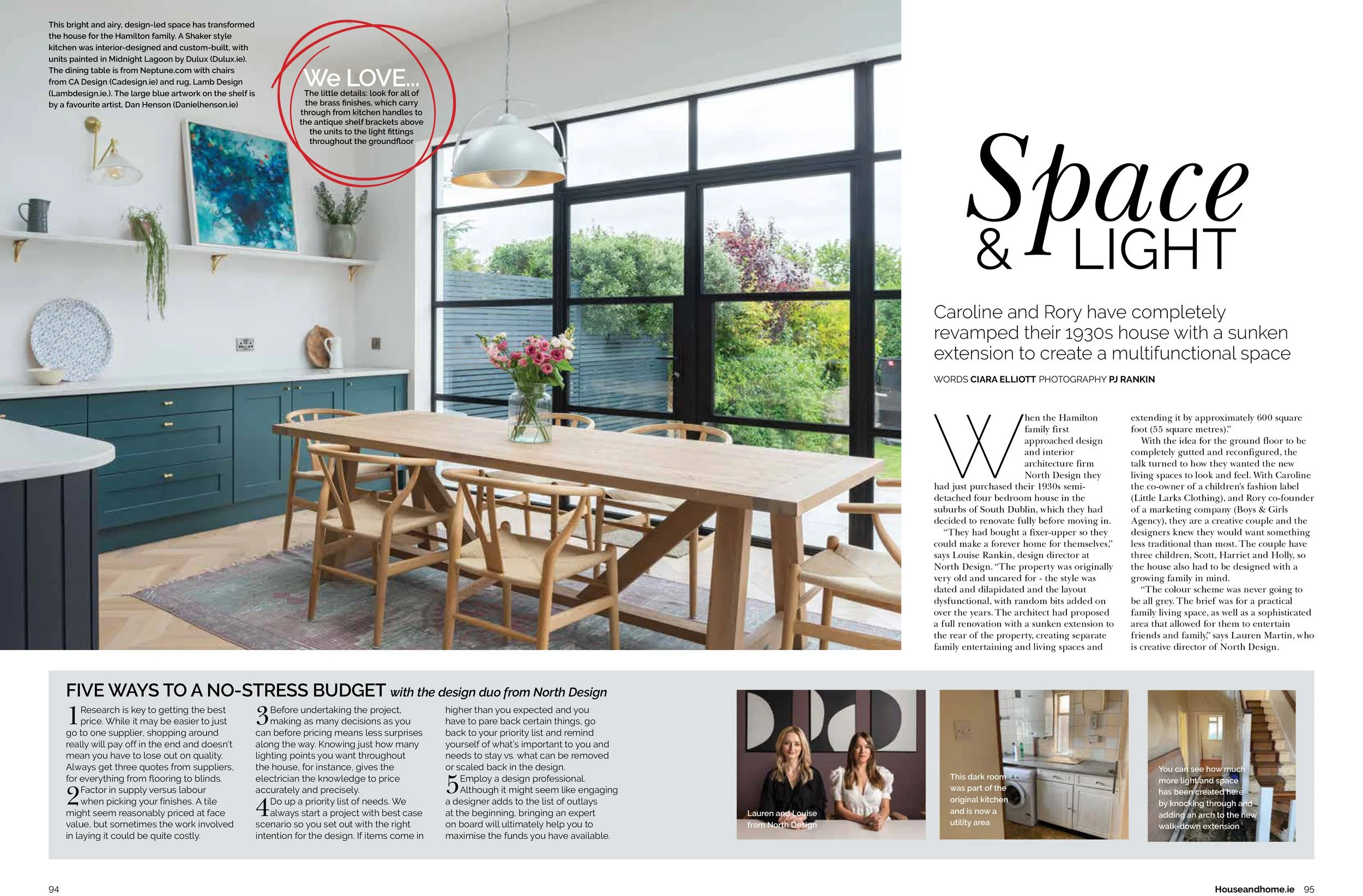 A bright, modern kitchen and dining area with large black-framed windows, a wooden dining table with chairs, teal cabinetry, and decorative shelves with artwork and plants.
