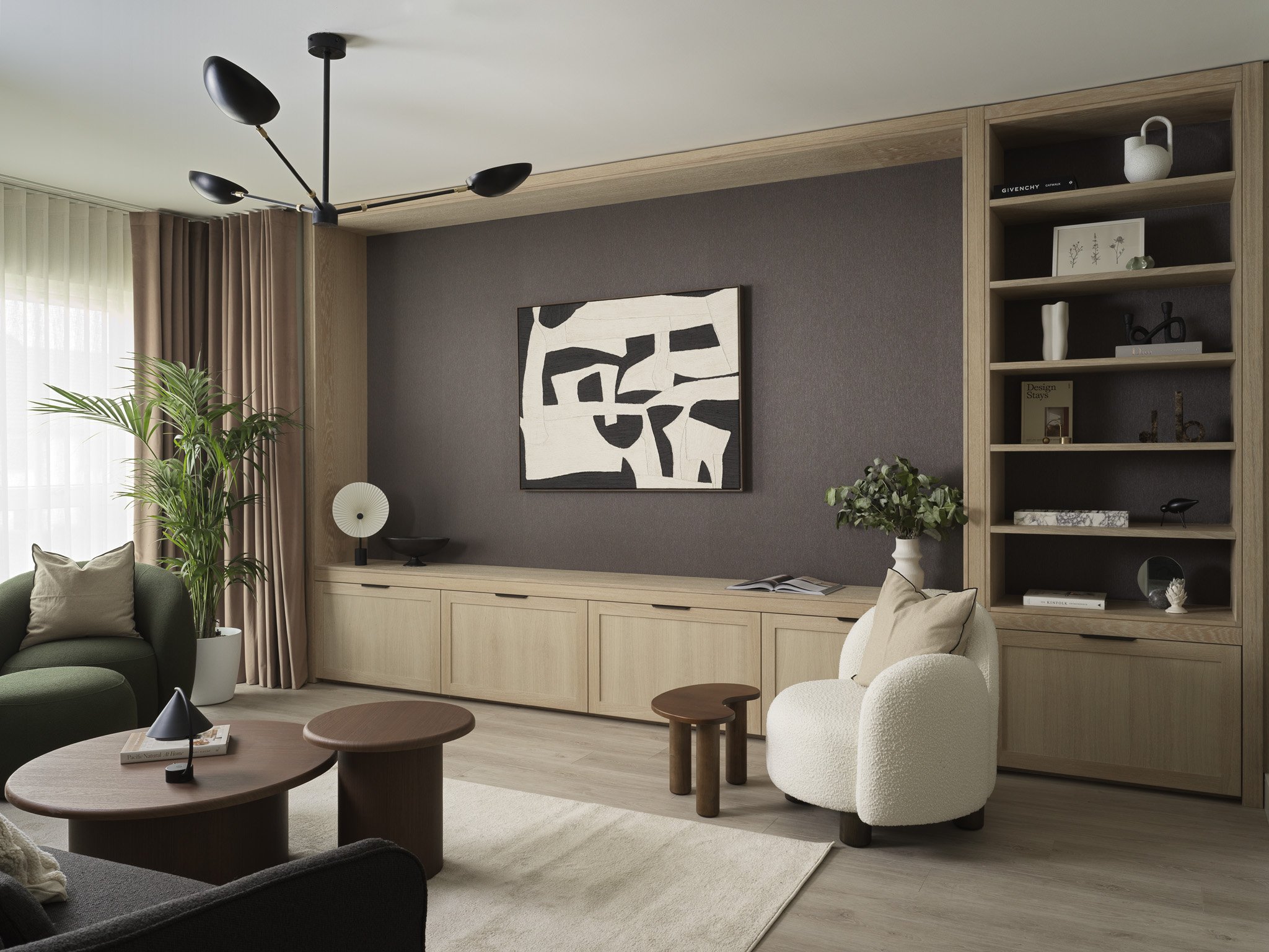 Modern living room with a beige and brown color scheme, featuring a round wooden coffee table, a white armchair with pillows, a sectional sofa, a tall potted plant, abstract artwork, built-in shelving, and curtains on a large window.