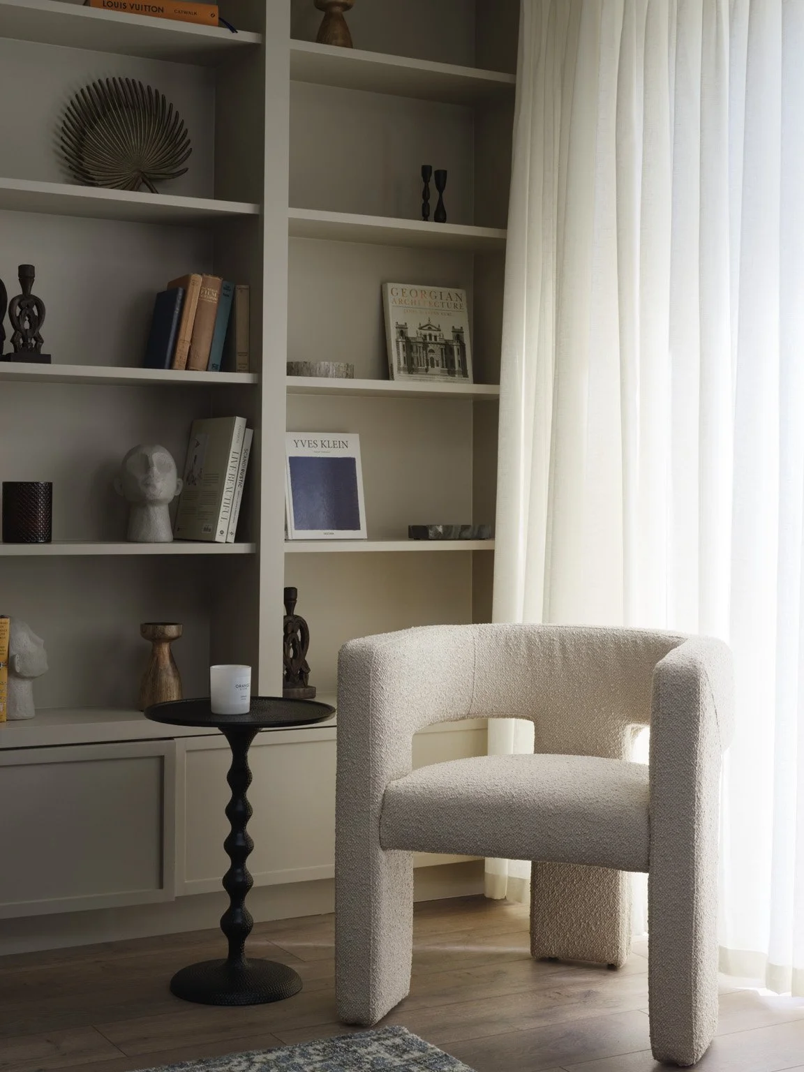 A cozy corner with white bookshelves, a plush beige armchair, a small round black side table with a white candle, and cream-colored curtains allowing natural light into the room.