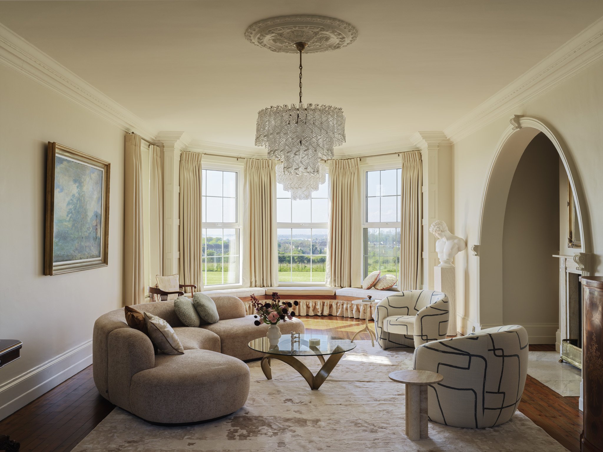 Elegant living room with large bay windows, beige curtains, a chandelier, a beige curved sofa, two patterned armchairs, a glass coffee table with a flower vase, a portrait on the wall, and a bust sculpture near an arched fireplace.