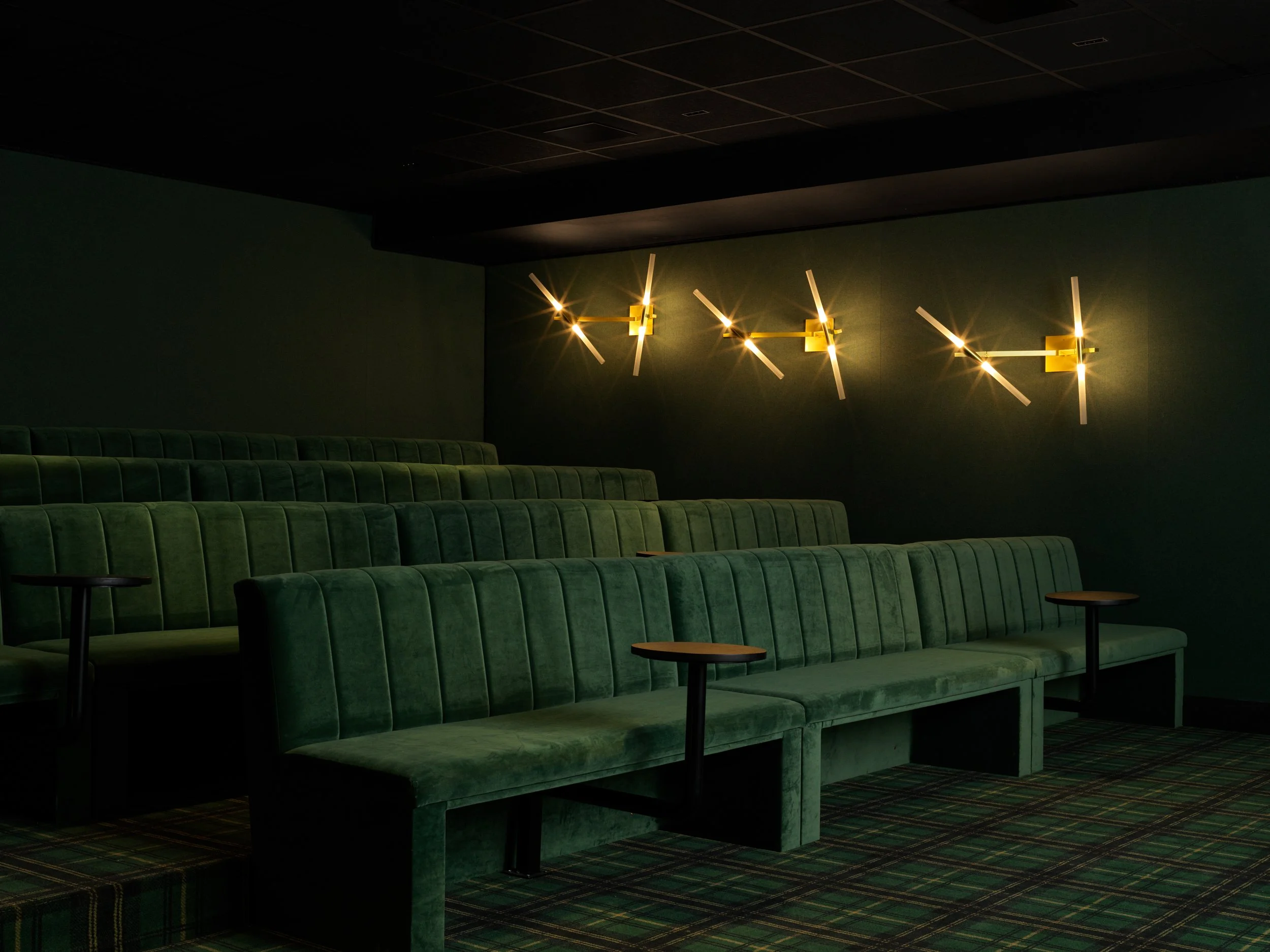 Green velvet theater seats arranged in rows with small wooden tablets on black metal stands, dimly lit with modern yellow wall sconces on a dark green wall and a patterned carpet.