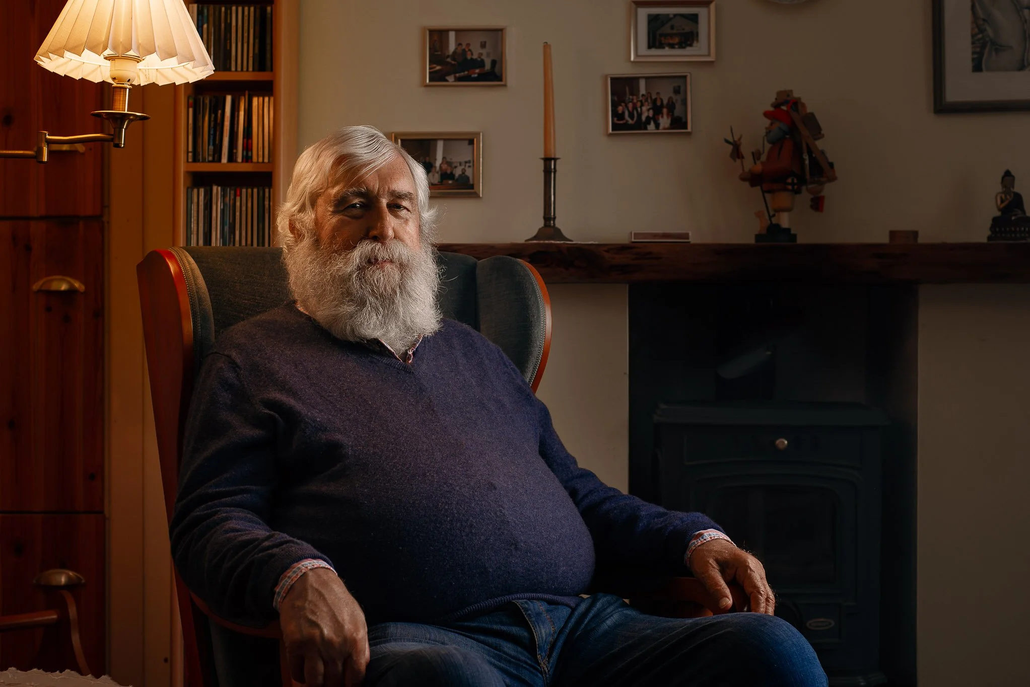 An elderly man with a white beard sitting in a dark green armchair in a cozy room with wooden walls, a bookshelf, framed photos, and decorative items in the background.