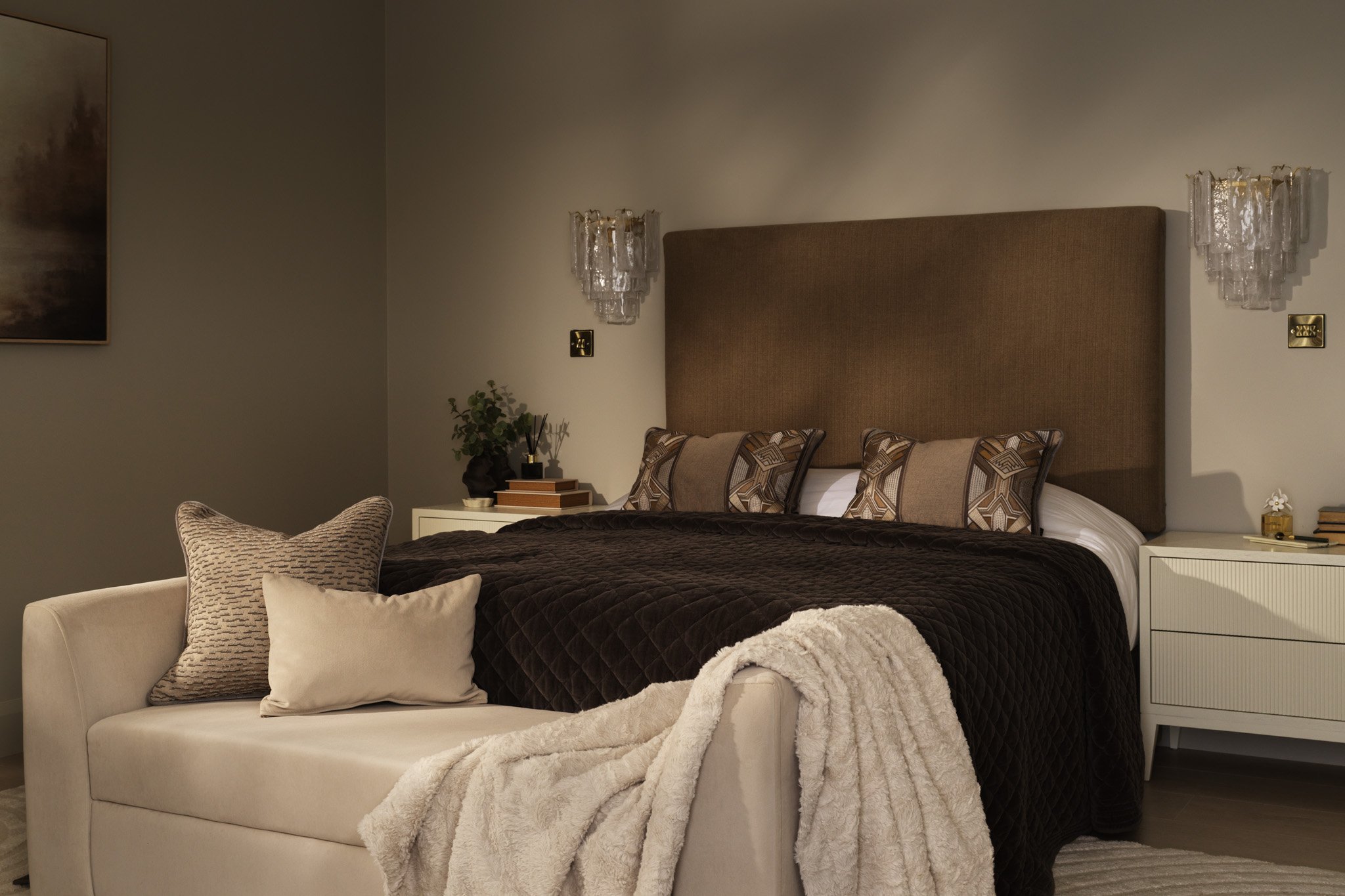 Elegant bedroom with a brown upholstered headboard, black quilted bedding, patterned pillows, crystal wall sconces, and a cream chaise lounge with a fluffy throw blanket.