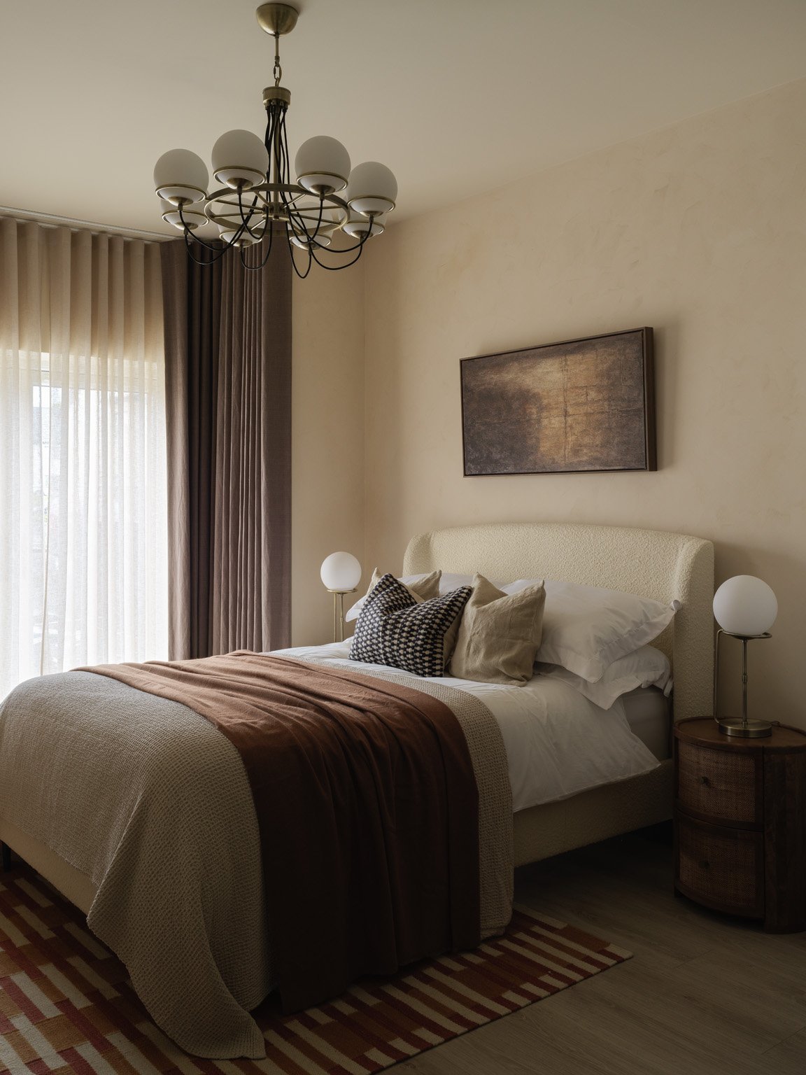 A bedroom with a bed, pillows, and a brown blanket. There are two round bedside lamps, a hanging chandelier, curtains covering a window, a framed artwork above the bed, and a striped rug on the floor.