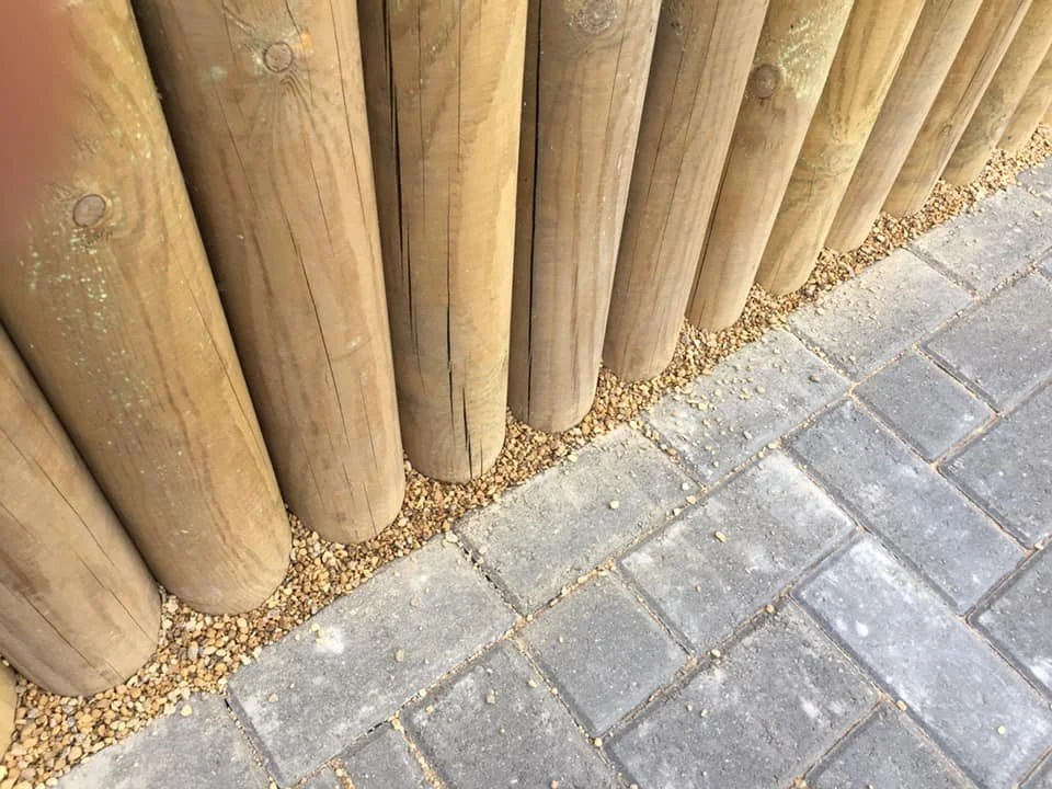 Close-up of a wooden fence with round logs, set on a bed of small gravel, next to a paved brick sidewalk.